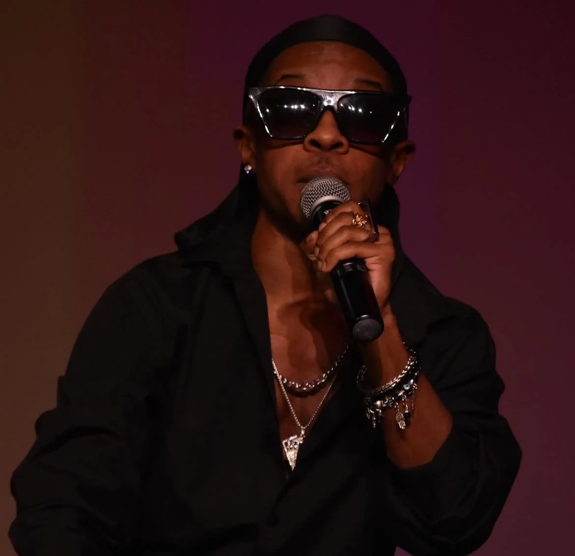 A person wearing dark sunglasses and a black outfit, holding a microphone, performing on stage with a dark background.