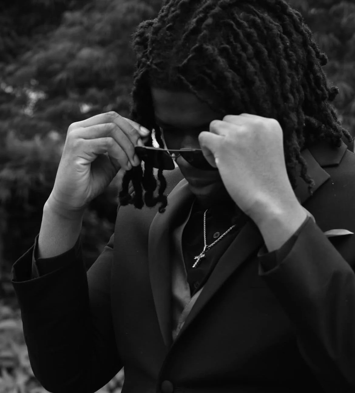 A person with shoulder-length dreadlocks adjusting sunglasses, wearing a blazer and a chain with a cross. The background is blurred with trees.