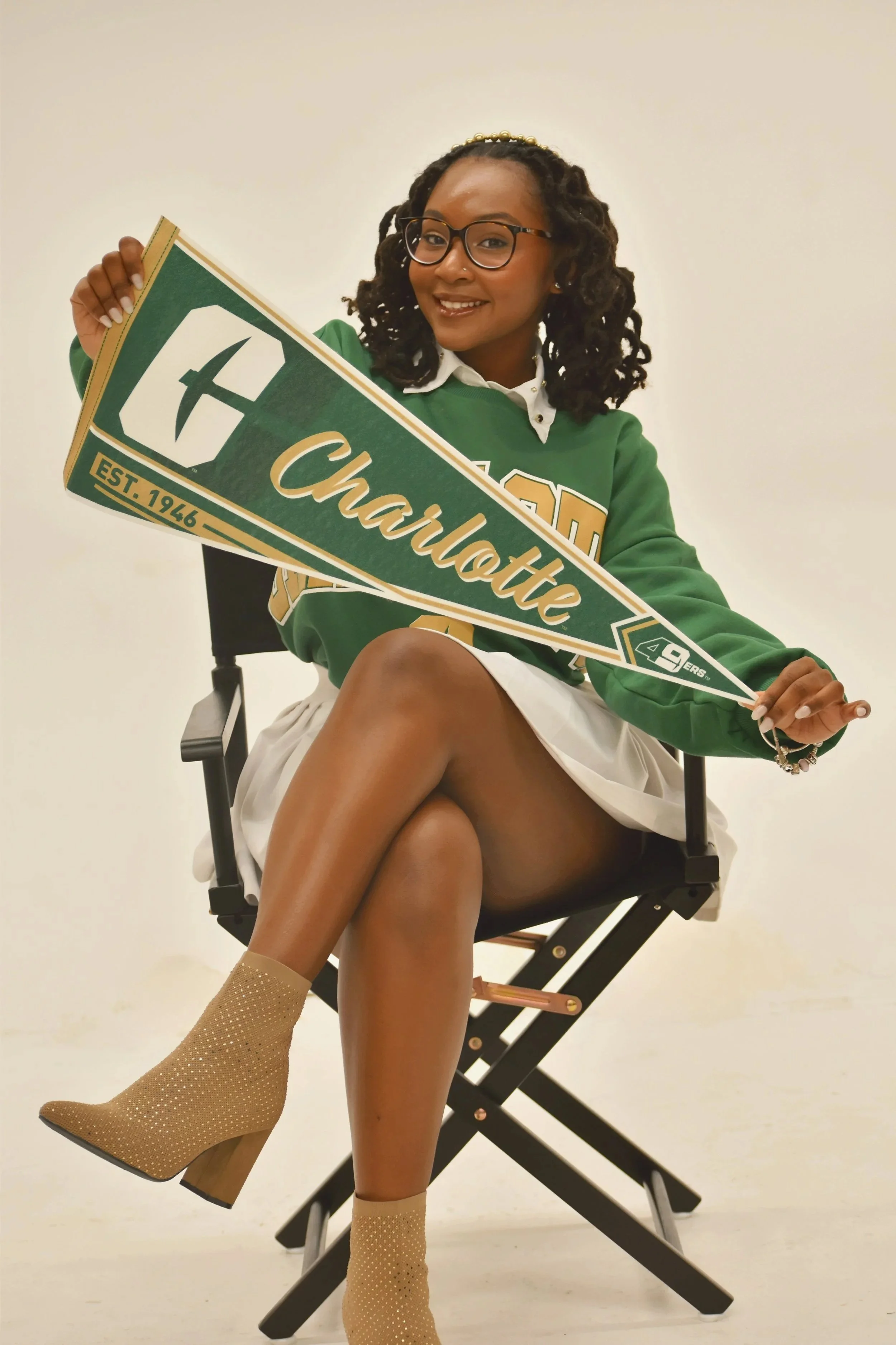 Young woman sitting on a director's chair, holding a green and white Charlotte Hornets pennant, wearing a green Charlotte sweatshirt, white skirt, and tan heeled ankle boots, smiling at the camera.