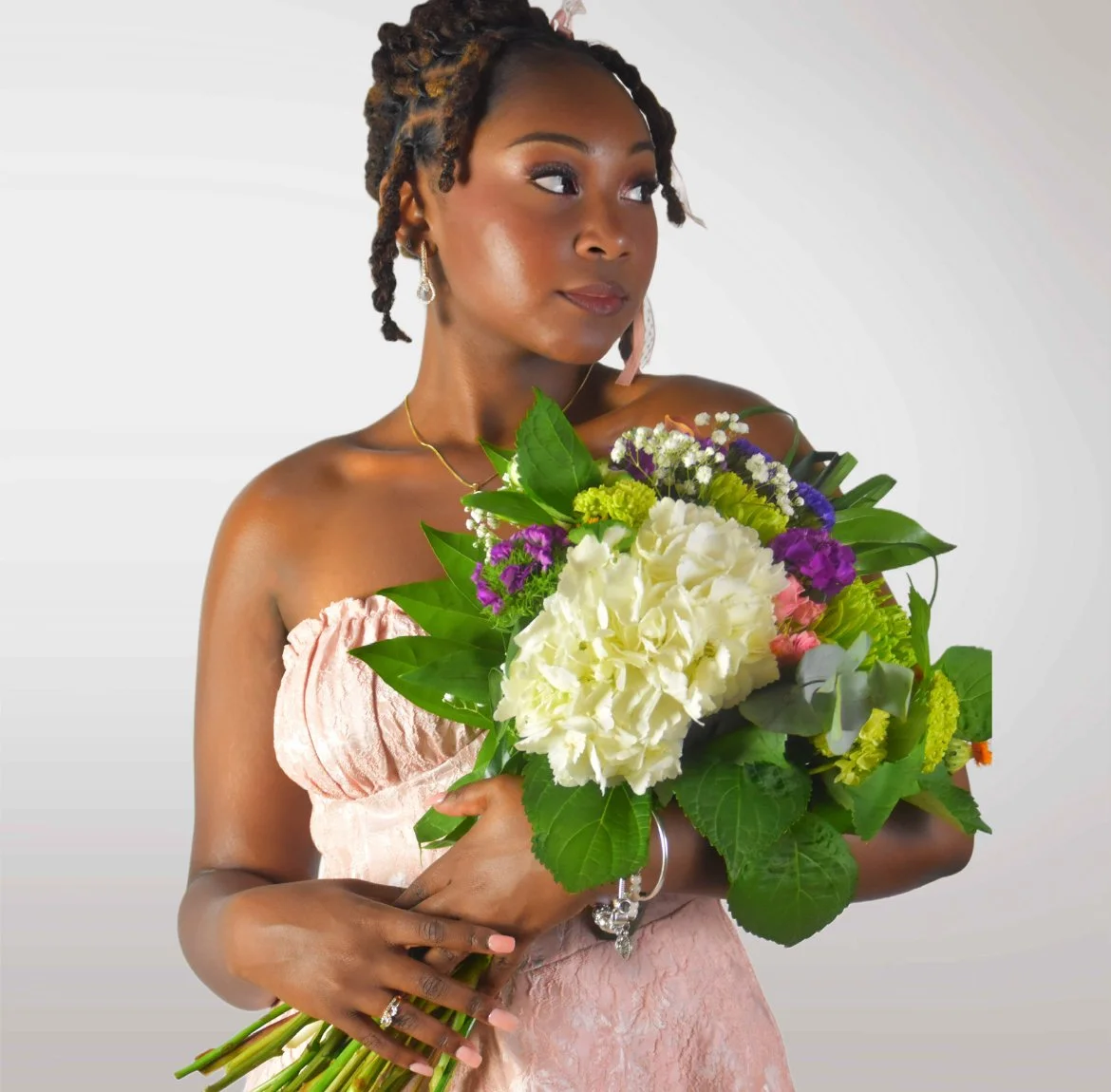 A woman with styled hair wearing a pink off-shoulder dress, holding a large colorful bouquet of flowers, looking to her right, against a plain grey background.