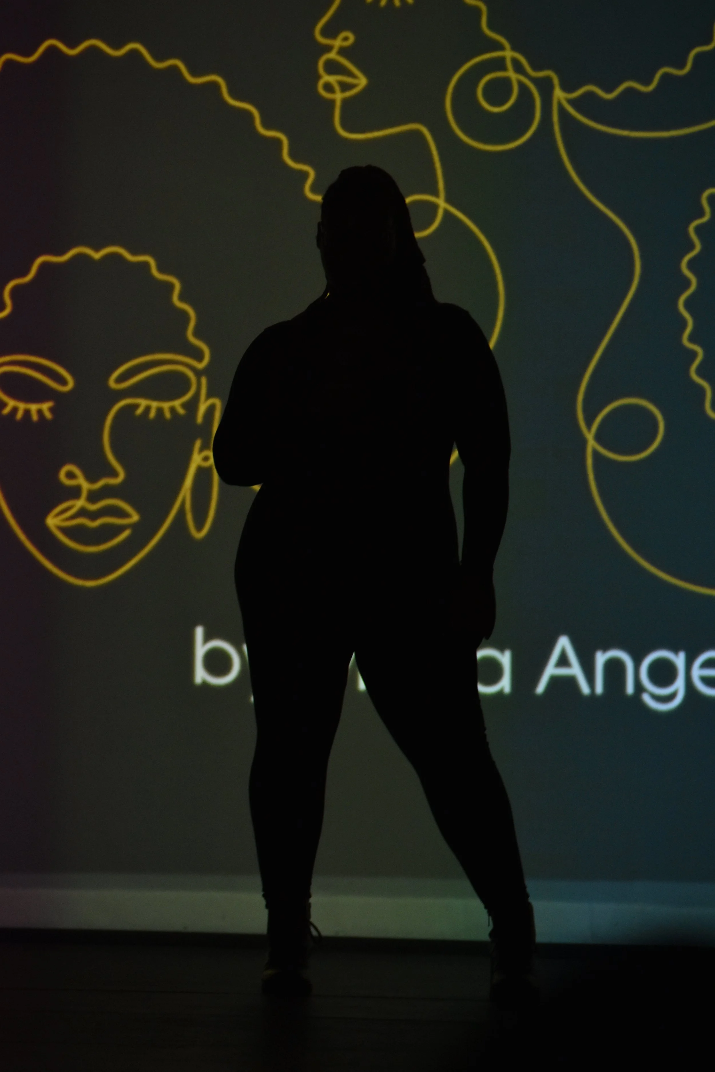 Silhouette of a woman standing in front of a wall with yellow line art portraits of women's faces and text.