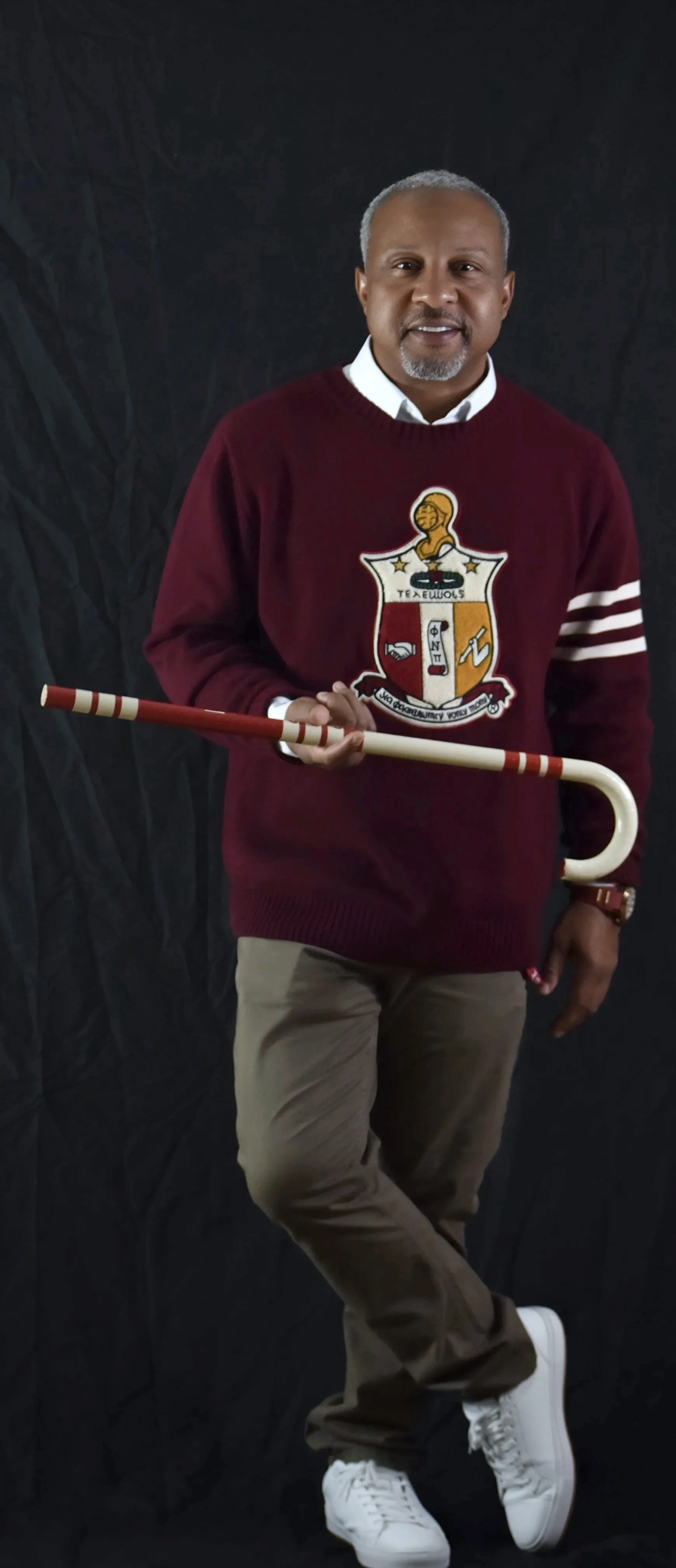 A man standing against a black backdrop, wearing a maroon sweater with a crest on it, holding a decorative cane, and dressed in khaki pants and white sneakers.