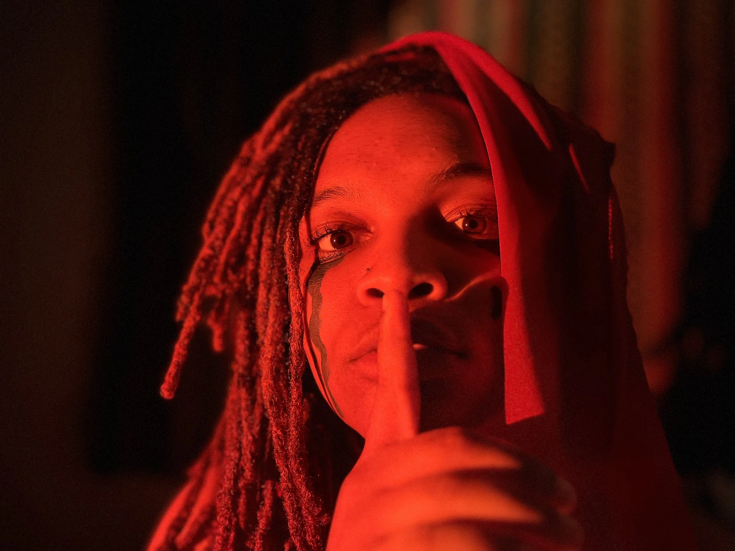 Close-up of a person with dreadlocks and face makeup illuminated by red light, holding a finger to lips in a shushing gesture.