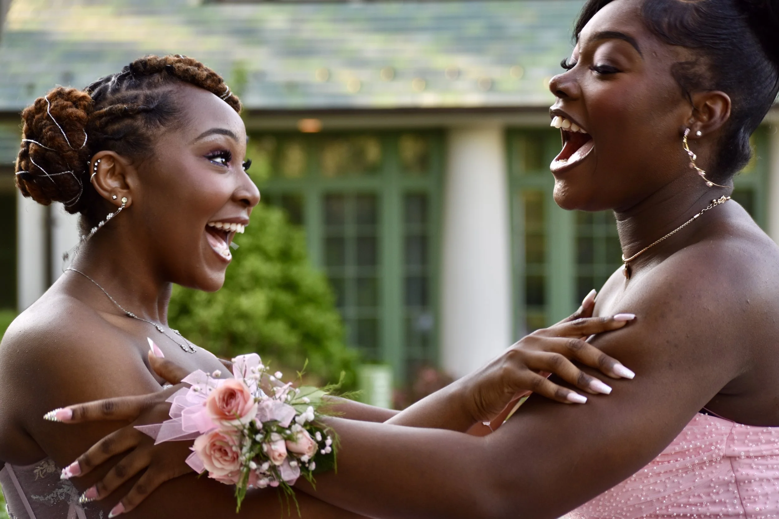 Two women in pink dresses are smiling and embracing each other outdoors, with a building and greenery in the background.