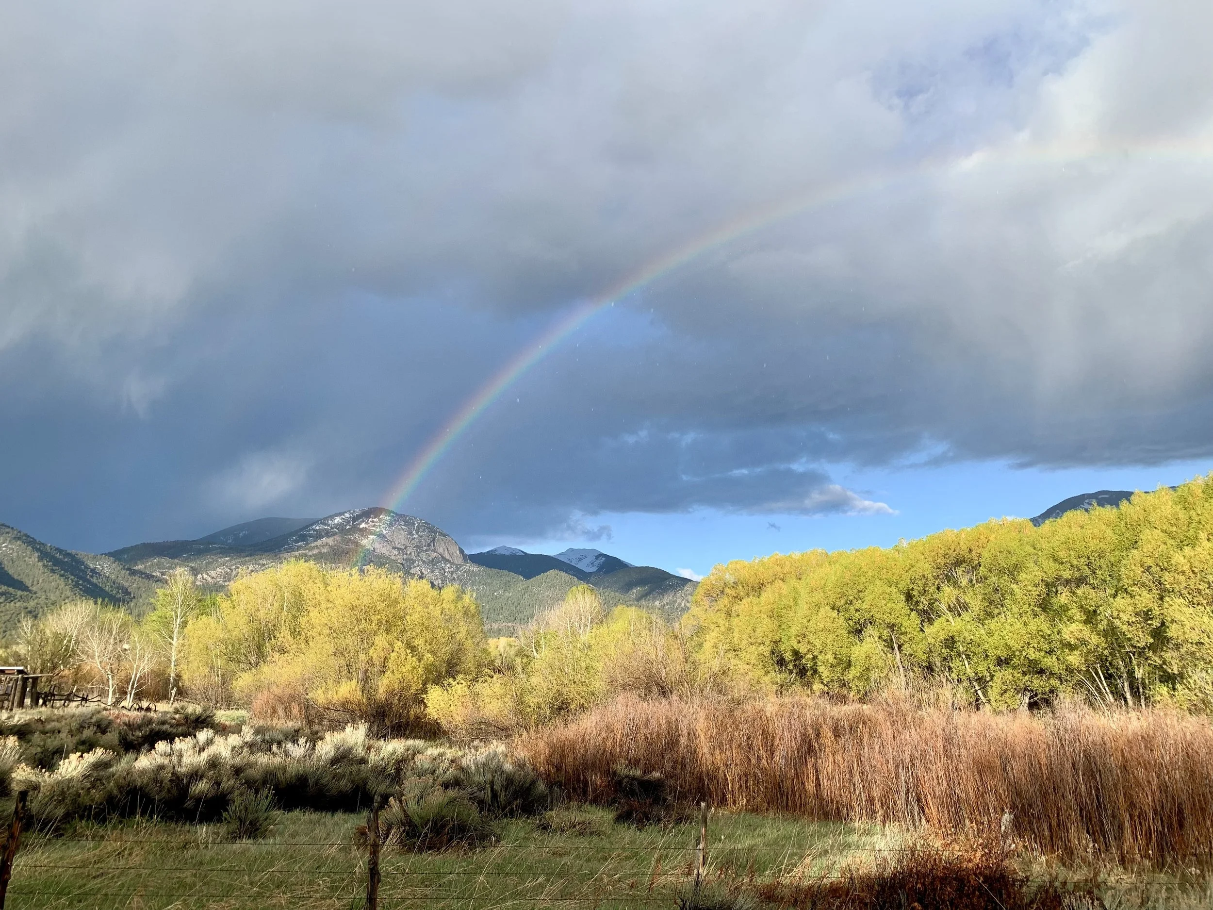 Panoramic view of a rainbow over mountain range with stormy clouds and bright sunlight illuminating the colorful foliage in a valley.