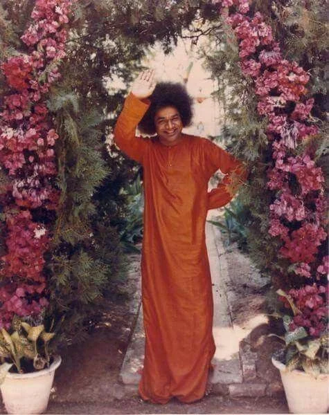 Sathya Sai Baba standing under an arch of flower in a garden setting.