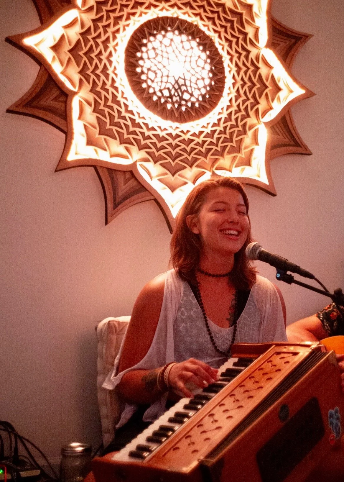 A woman smiling and playing a keyboard in front of a microphone, with a decorative, illuminated, star-shaped ceiling light behind her.