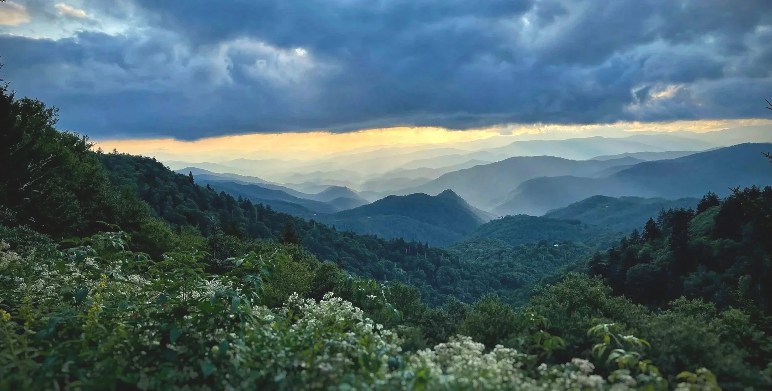 A scenic view of green, forested mountains under a dramatic sky with dark clouds and rays of sunlight breaking through.