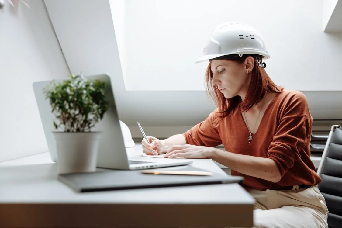 Woman in a hart hat working behind a computer and writing a note