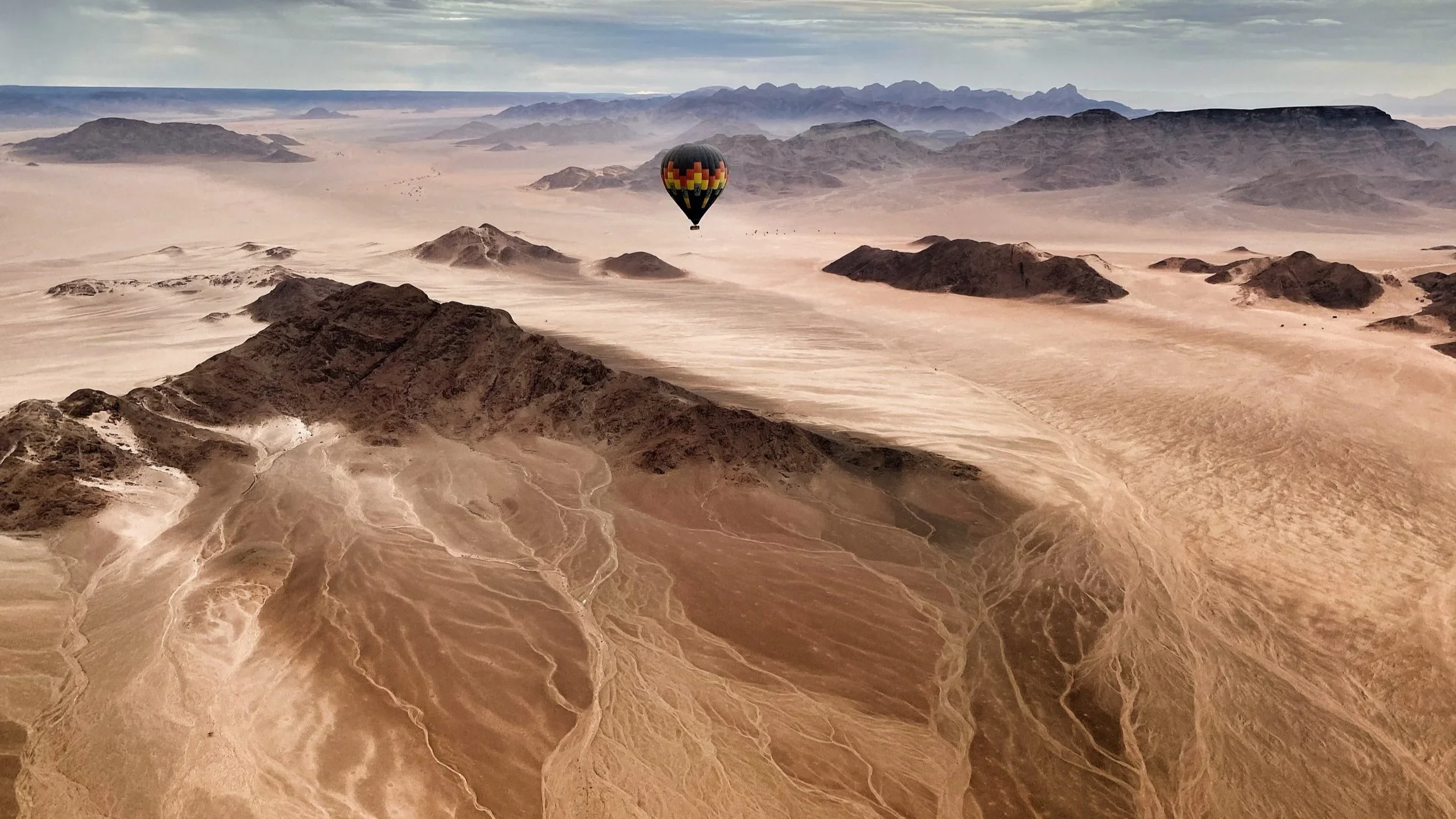 A hot air balloon floating over a desert landscape with sandy dunes and rugged mountains in the background.