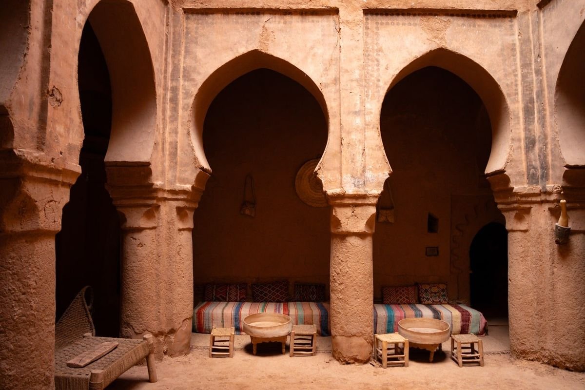 Interior of a traditional mud-brick structure with arched doorways, cushions on the floor, and small wooden tables, suggesting a Middle Eastern or North African setting.