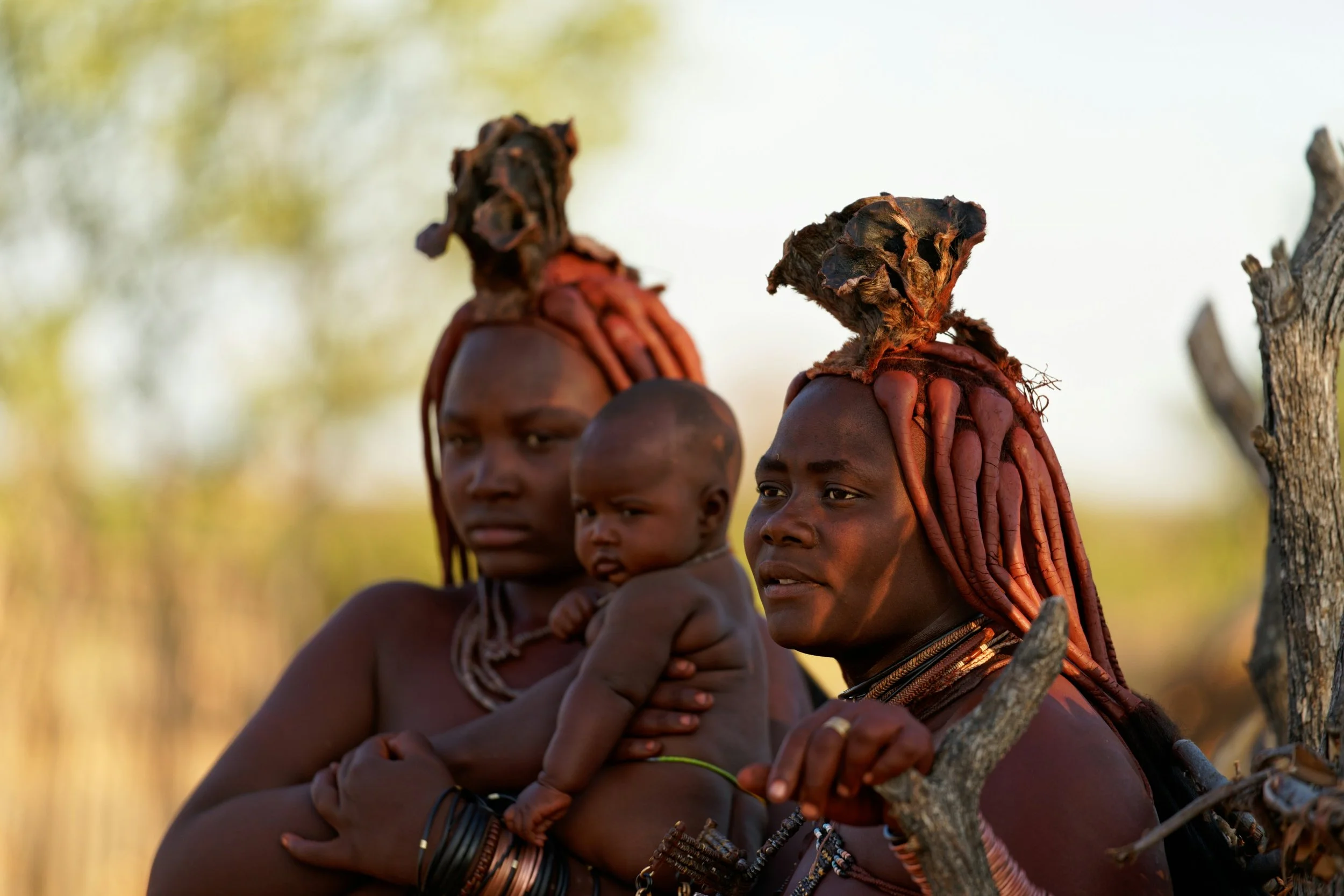 Tribal women from the Hamar ethnic group in Ethiopia, with traditional hairstyles and jewelry, one holding a child, standing outdoors in a natural setting.