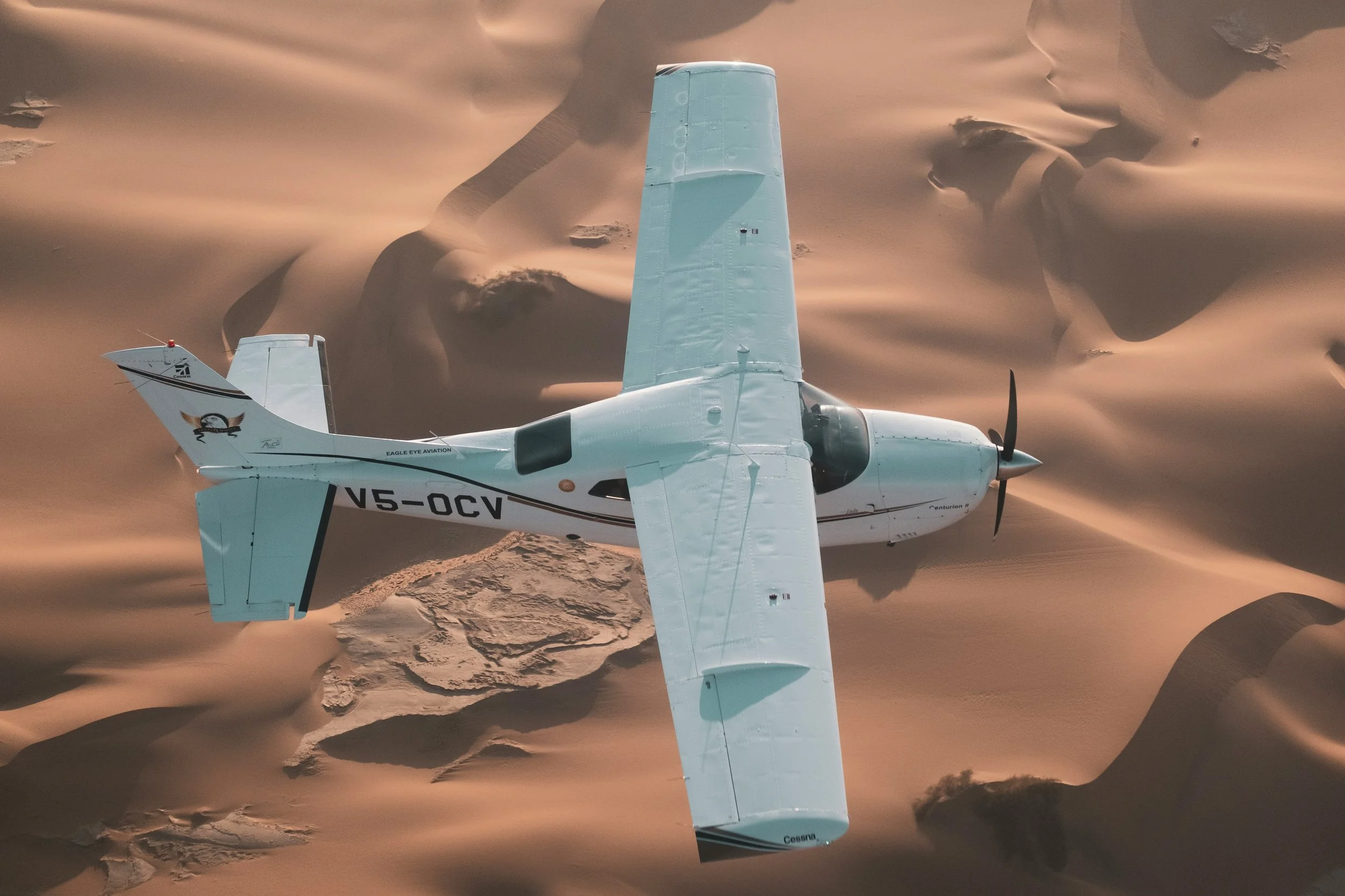 A small light aircraft flying over sand dunes in a desert landscape.