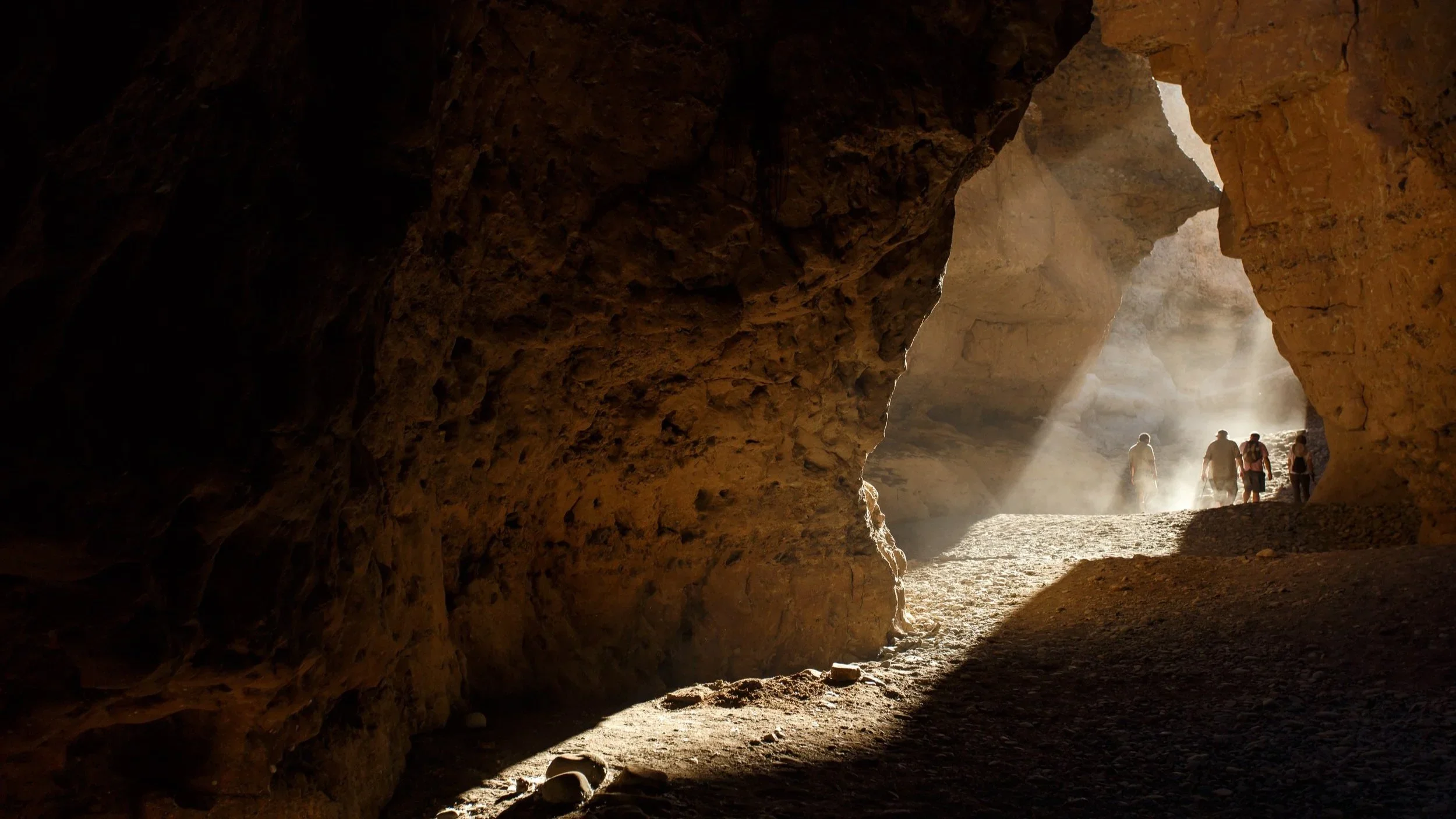 A view from inside a cave looking out toward the sunlight. Four people are walking near the cave opening, with rays of light shining through dust in the air.