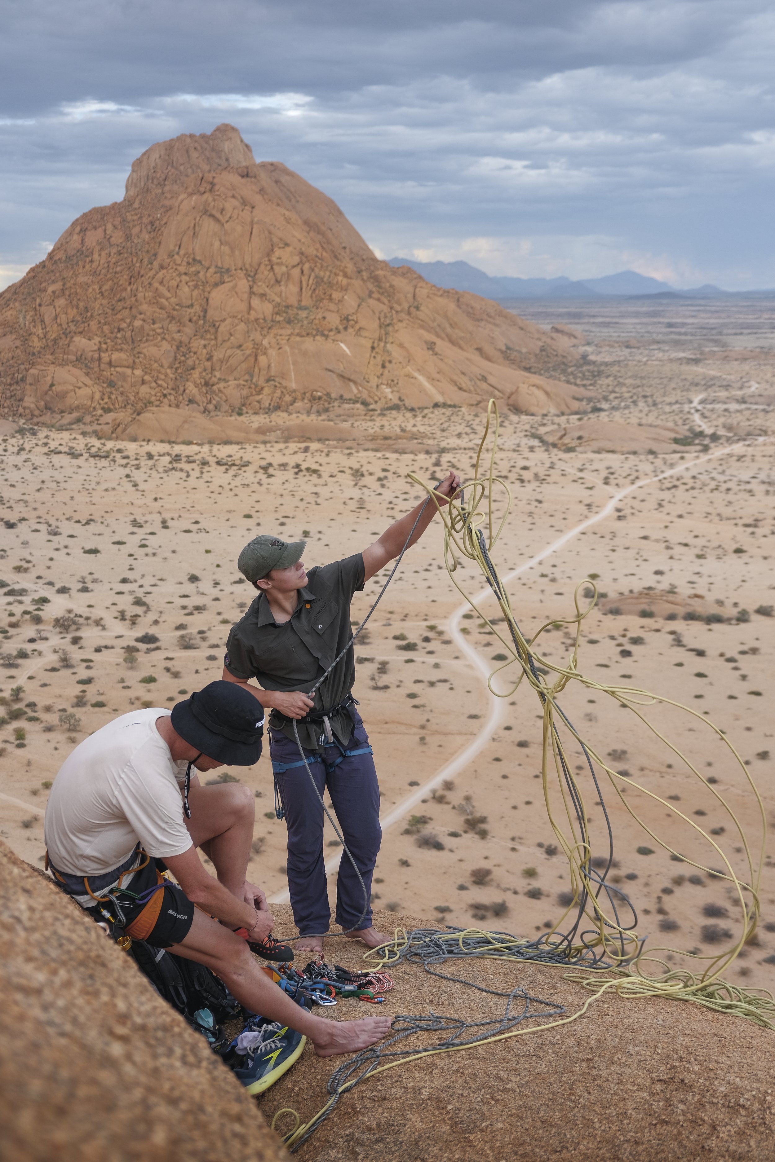Two climbers preparing to descend a rock formation in a desert landscape during cloudy weather, with large rocky hills in the background.