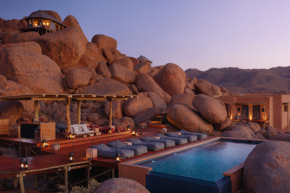 Luxury outdoor resort pool area with sun loungers, nestled among large rocks and boulders in a desert landscape at dusk.