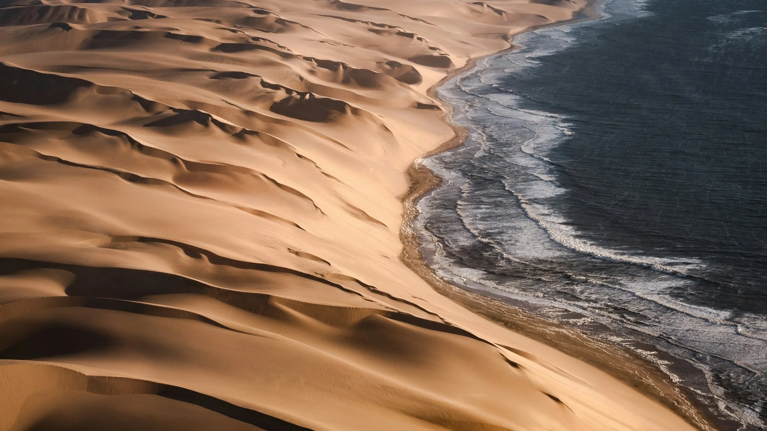 Aerial view of sand dunes meeting the ocean shoreline, with waves crashing onto the beach.