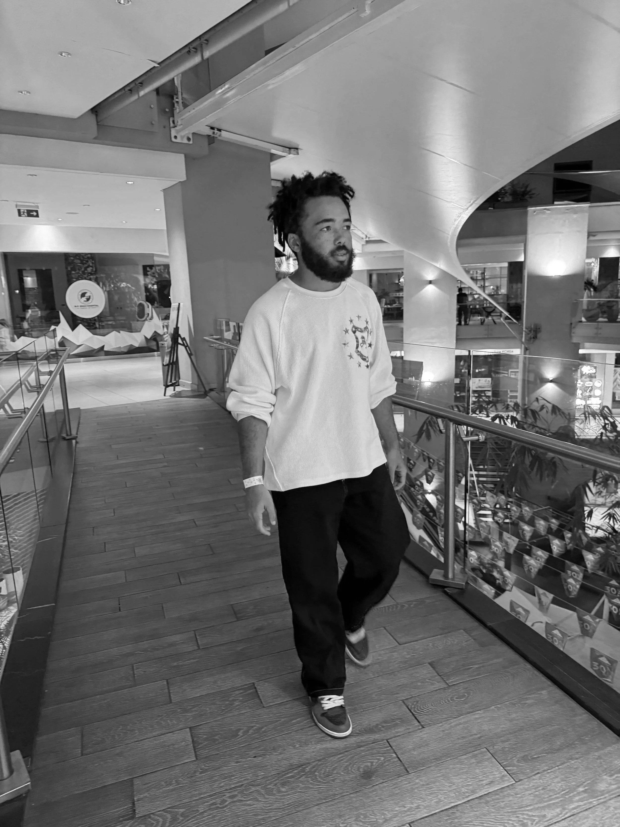 A man with dark brown hair and a beard wearing a white long sleeved top and black pants walking across a bridge.