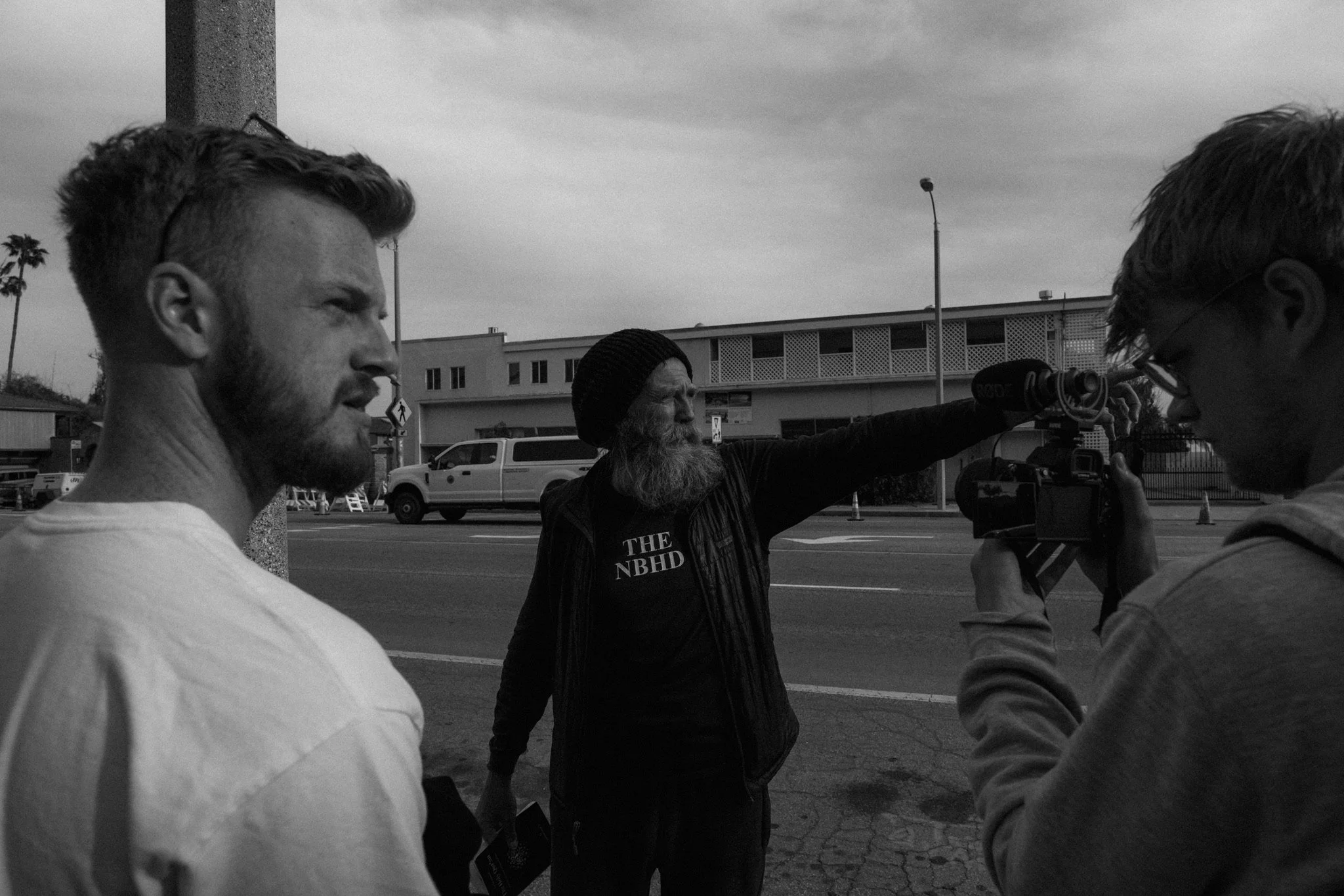 Two men looking to the right. One is wearing a beanie and a shirt that reads "THE NBHD" and is pointing to the right. The other is to the left of him, wearing a white t-shirt. On the right is a camera filming the man pointing. 