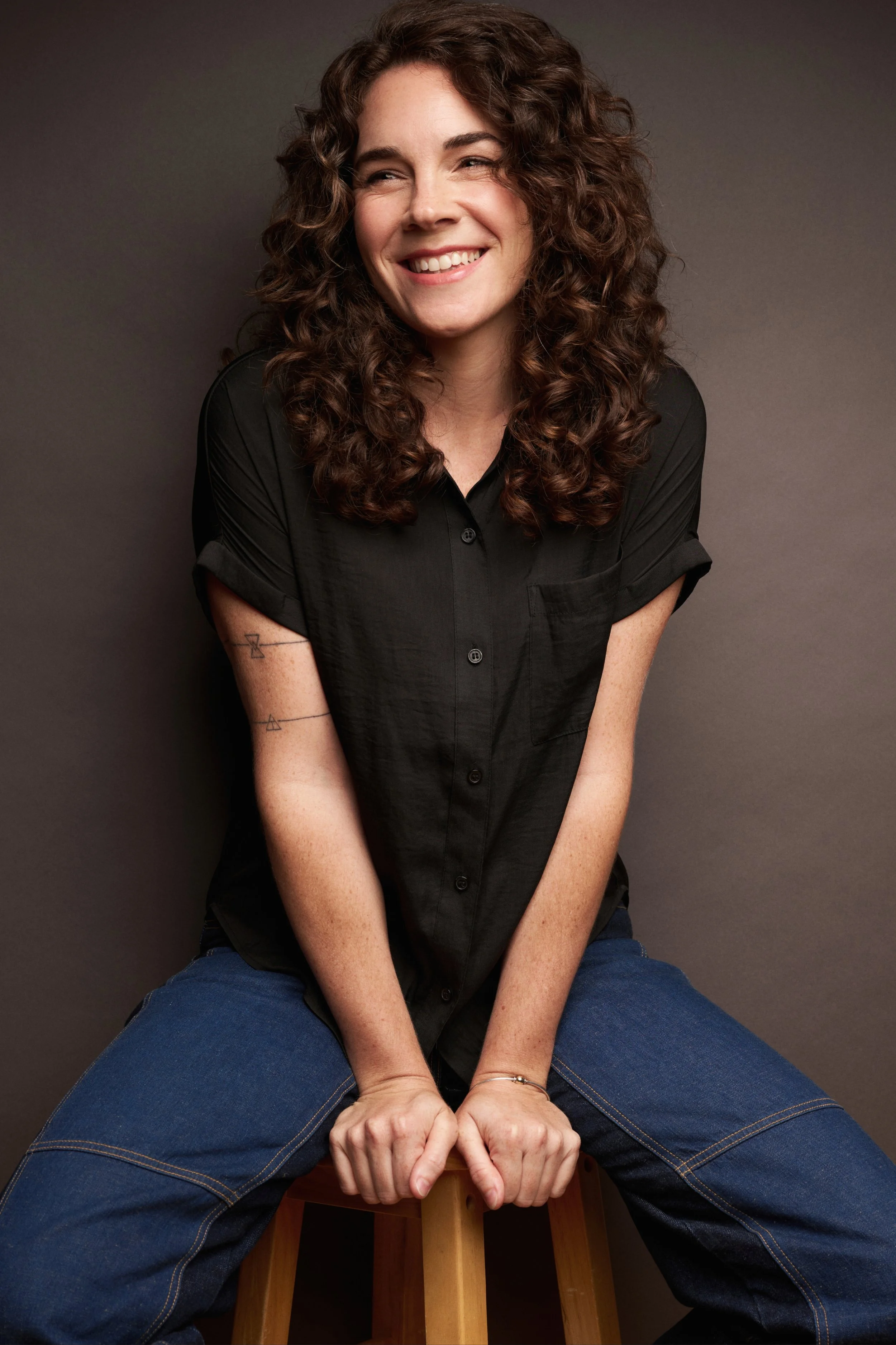 A woman with curly brown hair wearing a black shirt and blue jeans, sitting on a wooden stool, smiling and looking to the side against a gray background.