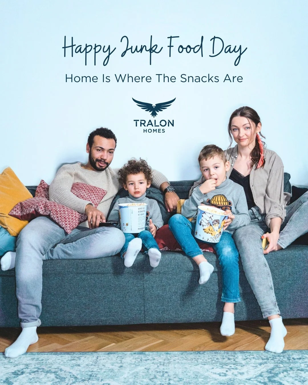A family of four sitting on a gray sofa with pillows, watching TV and eating popcorn at home, celebrating Junk Food Day.