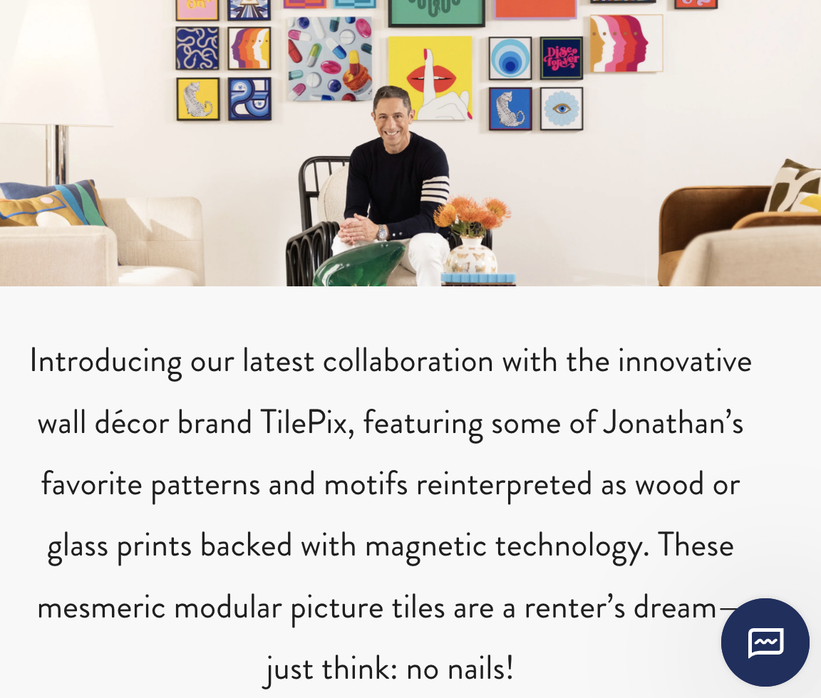 A man sitting in front of a colorful wall art display, smiling, with a table of flowers and decorative objects in front of him, in a bright, modern living room.