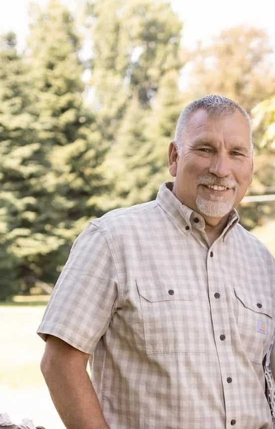 A smiling middle-aged man with short gray hair and a beard, wearing a beige plaid short-sleeve button-up shirt, standing outdoors in front of green trees.