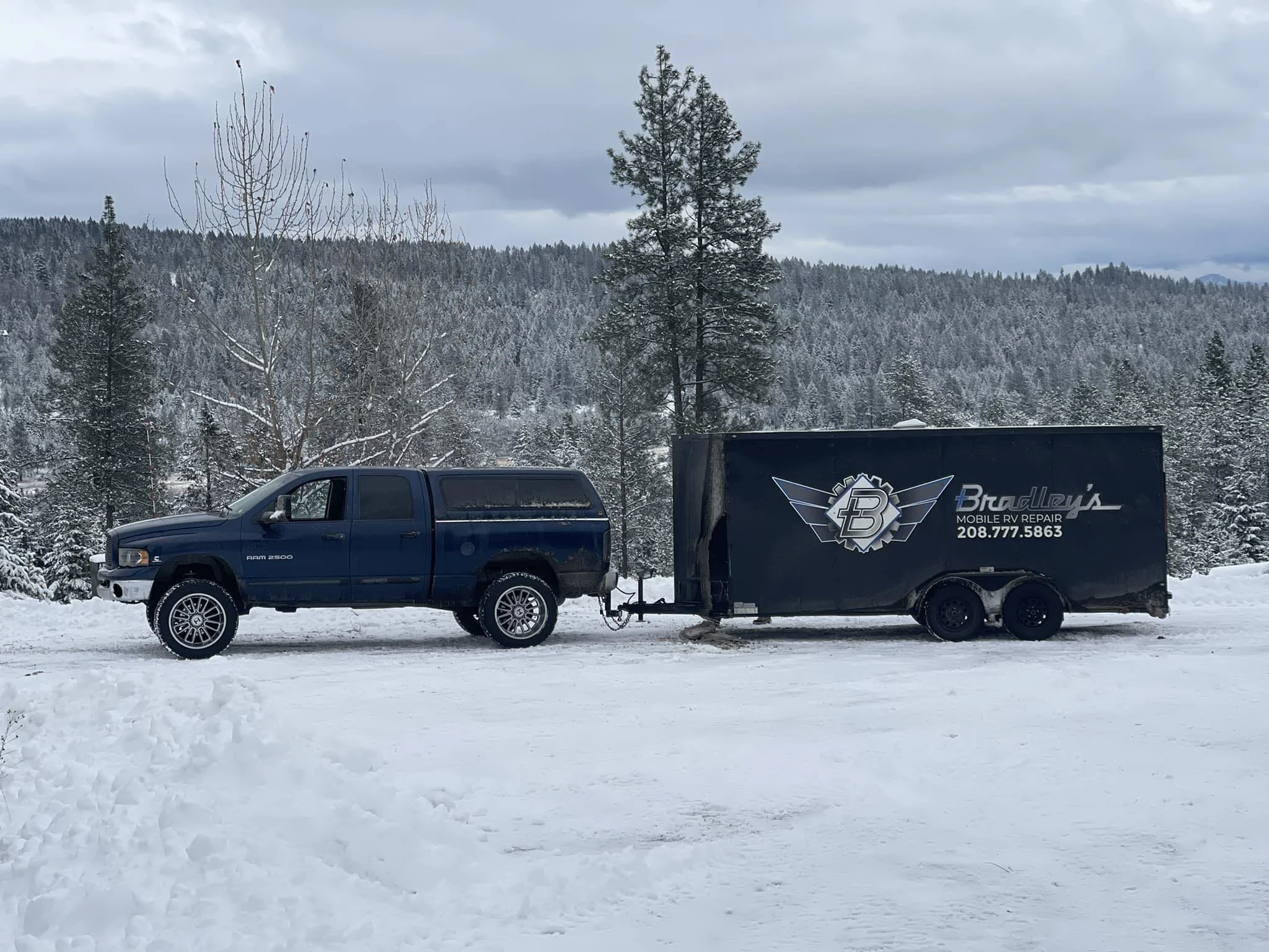 Blue pickup truck towing a black trailer with a logo and contact information, parked on snow-covered ground with trees and hilly landscape in the background.
