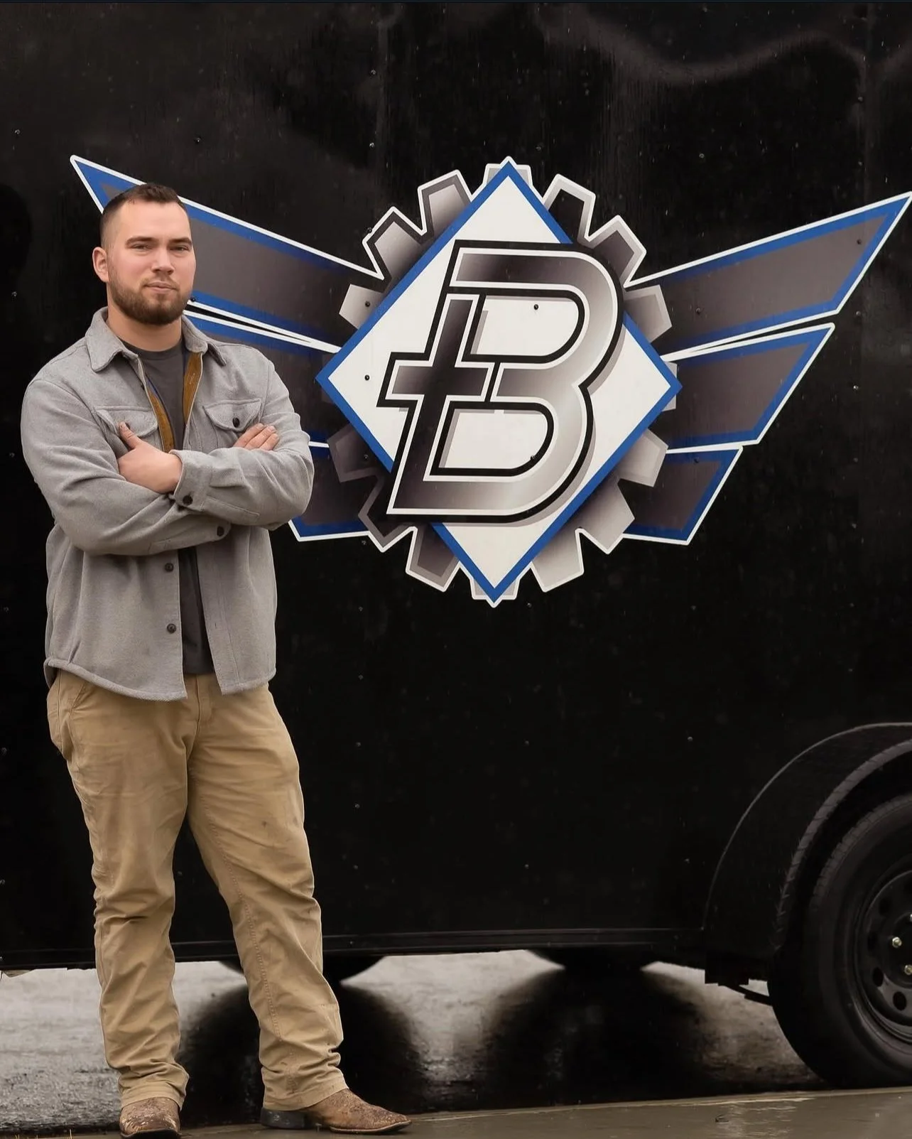 A man with a beard, wearing a gray jacket and beige pants, standing with arms crossed in front of a black vehicle with an emblem that has a large 'B' and a gear symbol.