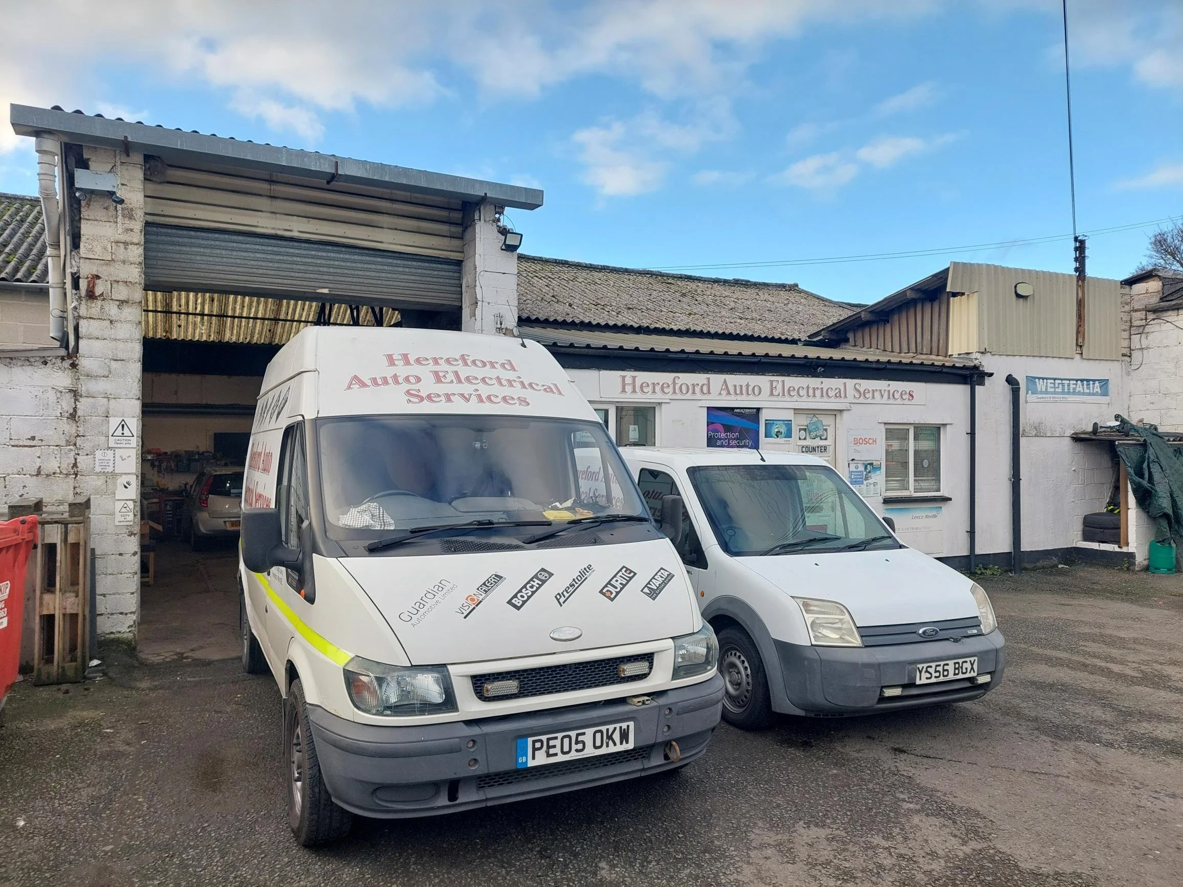 Outside of Hereford Auto Electrical Services with two white service vehicles parked in front, garage doors open revealing a car inside, building with signage and windows, blue sky with scattered clouds.
