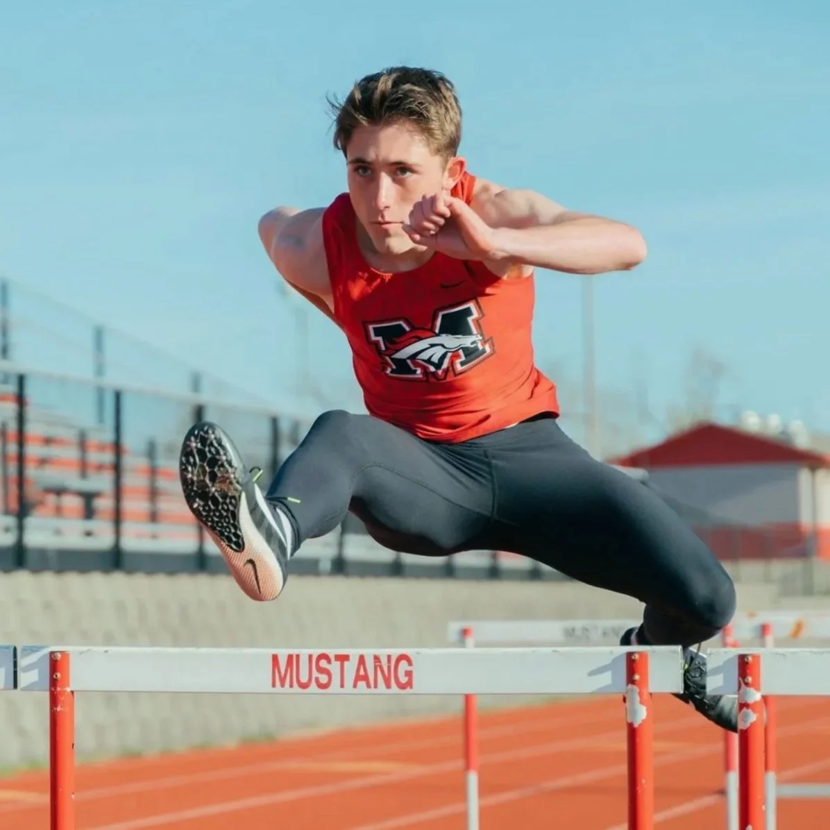 Male athlete in red shirt and black pants jumping over a hurdles on a track field outdoors.