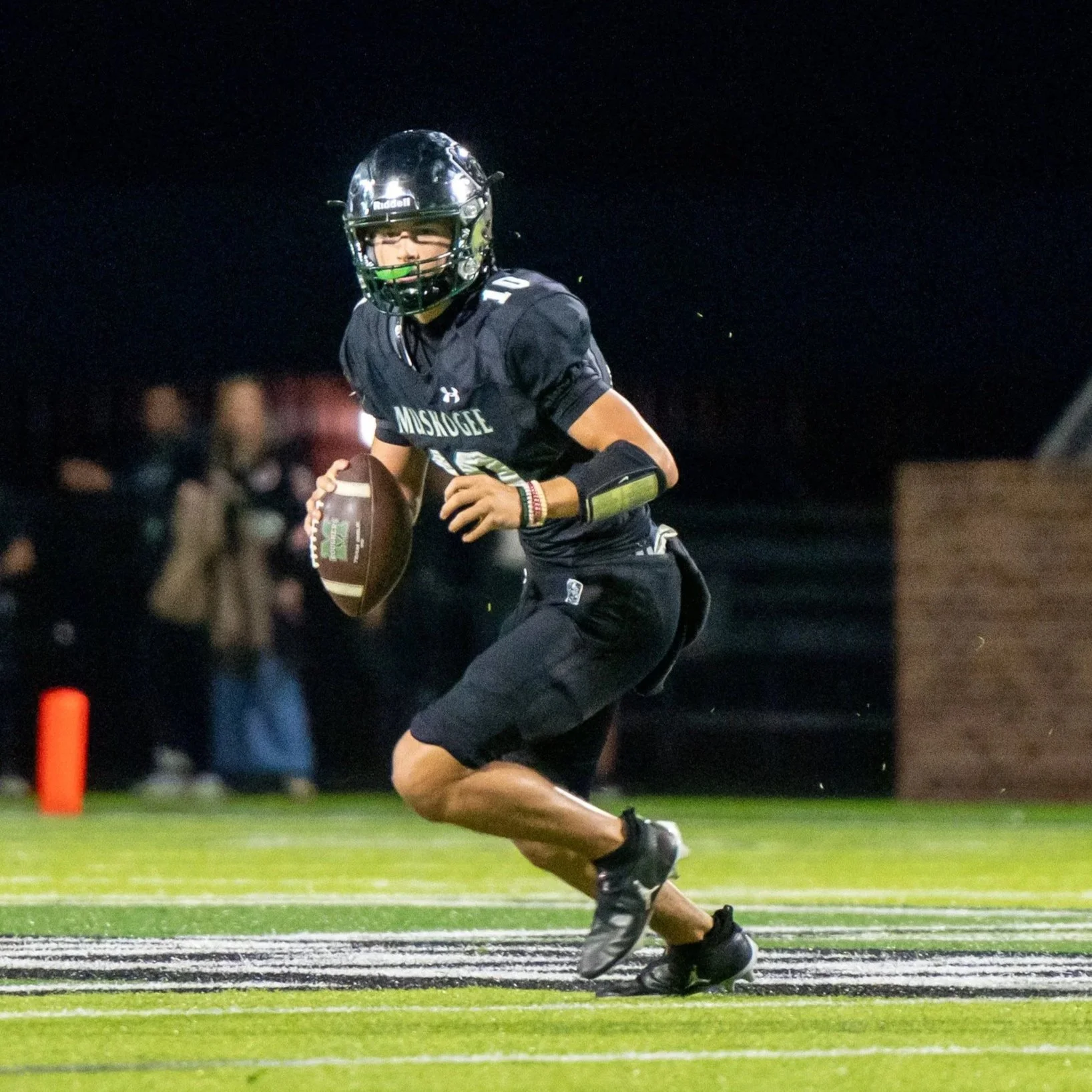 A football player in black uniform running with the ball on a field at night.