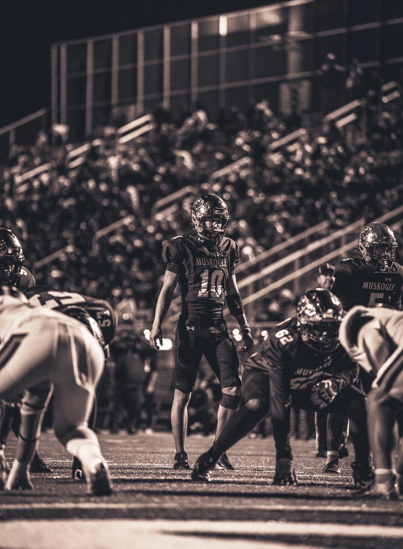 A black-and-white photo of a football game with players on the field, including one player standing in the center wearing a jersey with the number 10 and the word "Muskogee" on it. Other players are crouched in position, ready for play, with a crowd 
