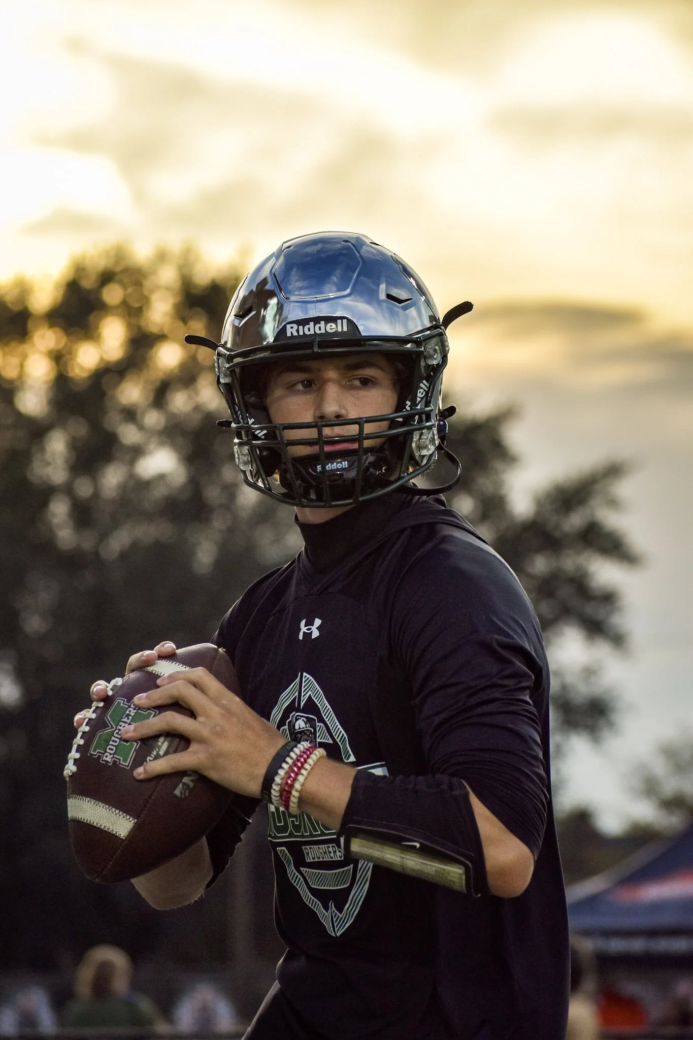 A young man wearing a football helmet and black sports shirt holding a football during a game at sunset.