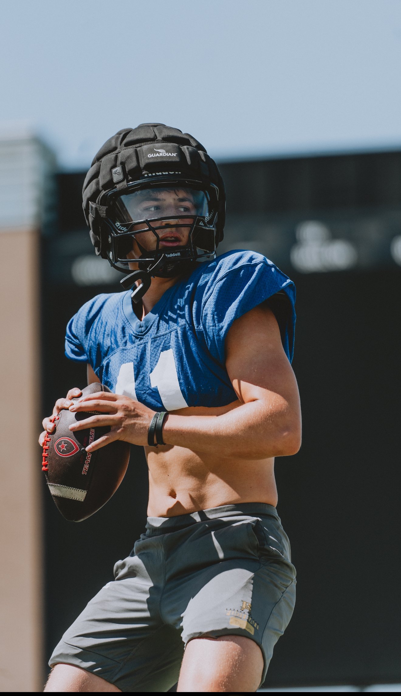 A young male football player in a blue jersey and gray shorts is holding a football during practice. He's wearing a black helmet with a clear face shield.