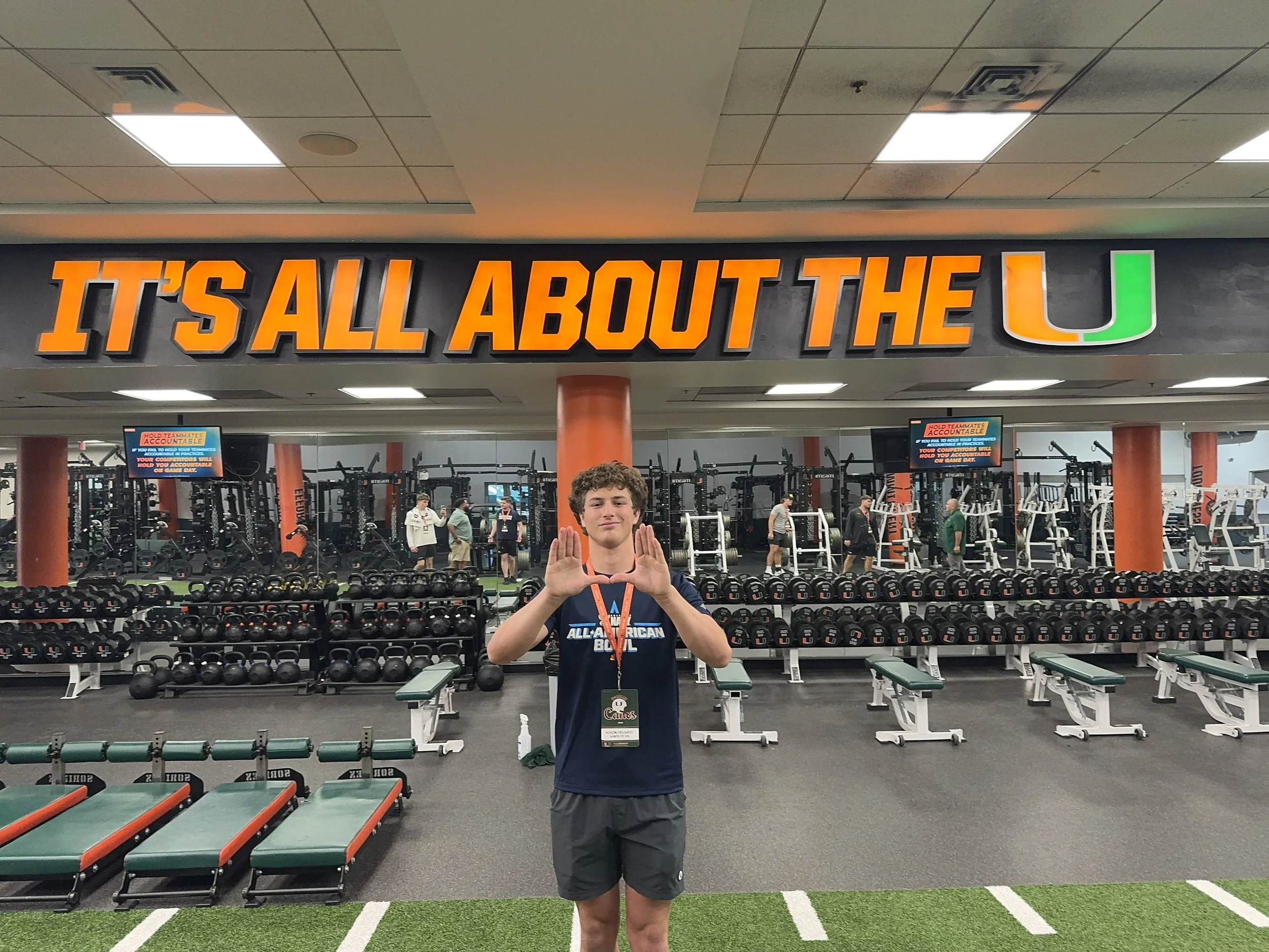 A young man standing in front of a gym with the sign 'IT'S ALL ABOUT THE U.' He is making a diamond shape with his hands and wearing a dark blue T-shirt with the words 'All-American Bowl' on it, and gray shorts.