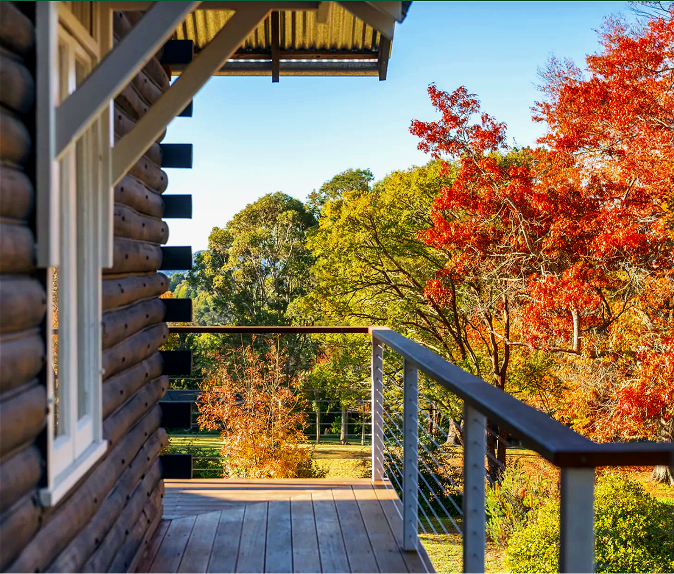 View from a wooden balcony with metal railing, overlooking colorful autumn trees with red, orange, yellow, and green leaves, under a clear blue sky.