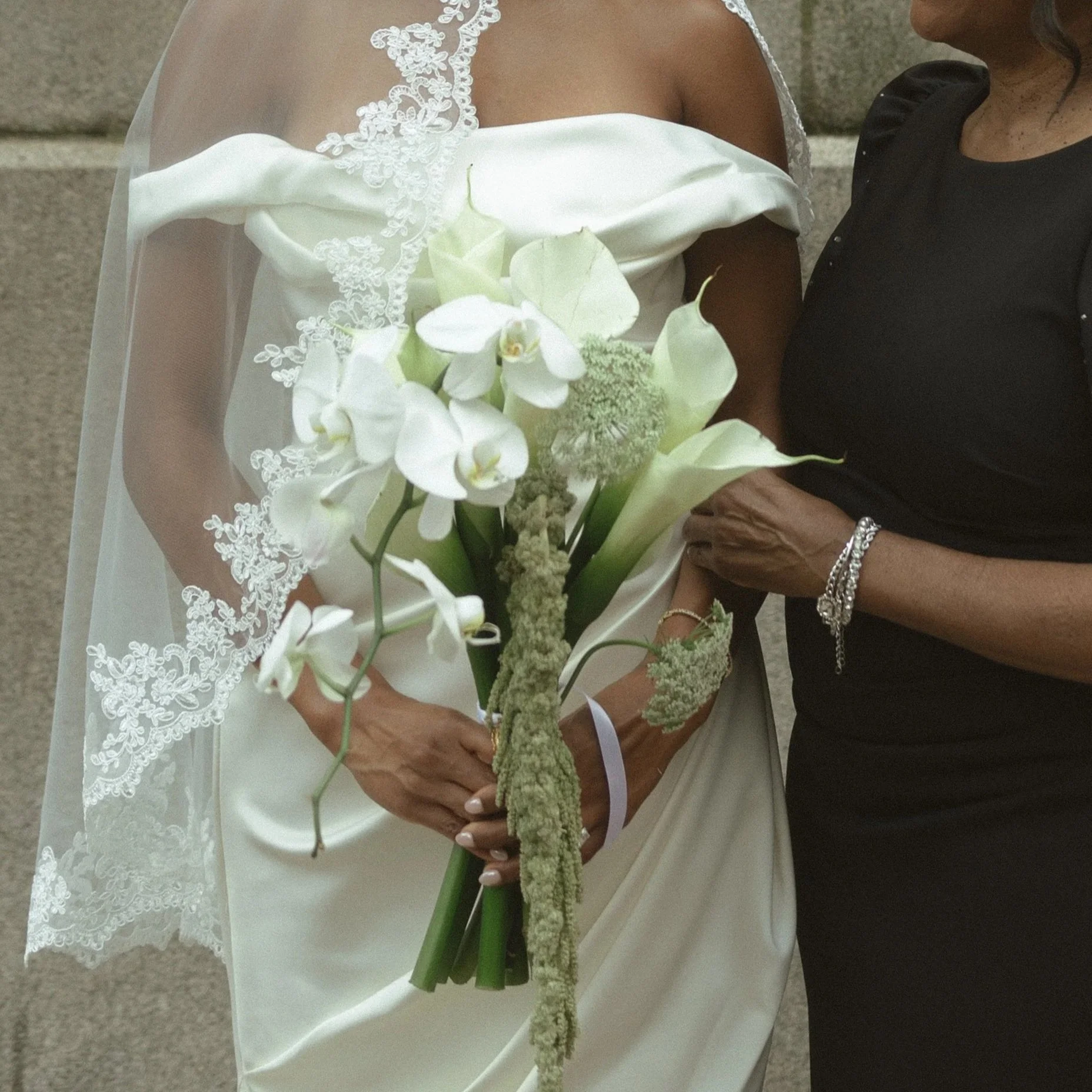 Close-up of a bride in a wedding dress holding a bouquet of white flowers, with another woman standing beside her.