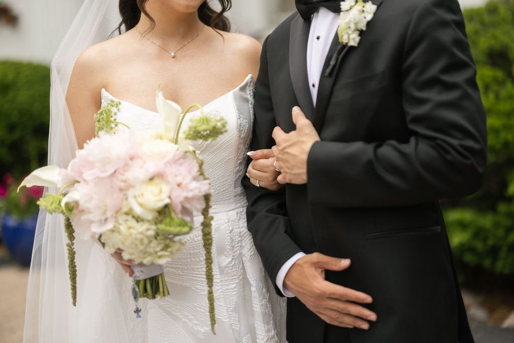Bride in a white wedding dress holding a bouquet of pink and white flowers, standing beside a groom in a black tuxedo with a white shirt and black bow tie, outdoors with greenery in the background.
