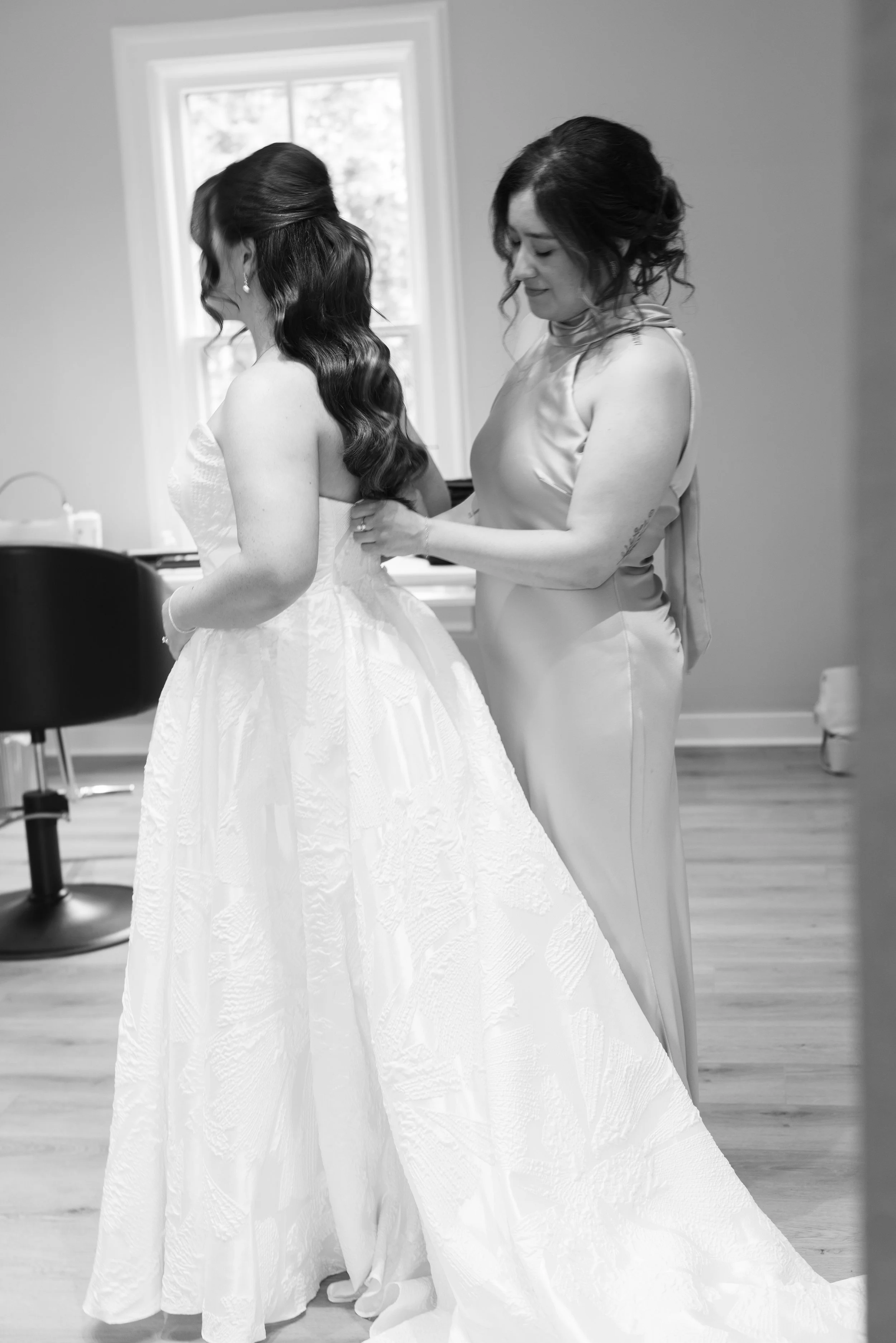 A bride getting help with her wedding dress from a woman in a satin gown, in a room with a window behind them.