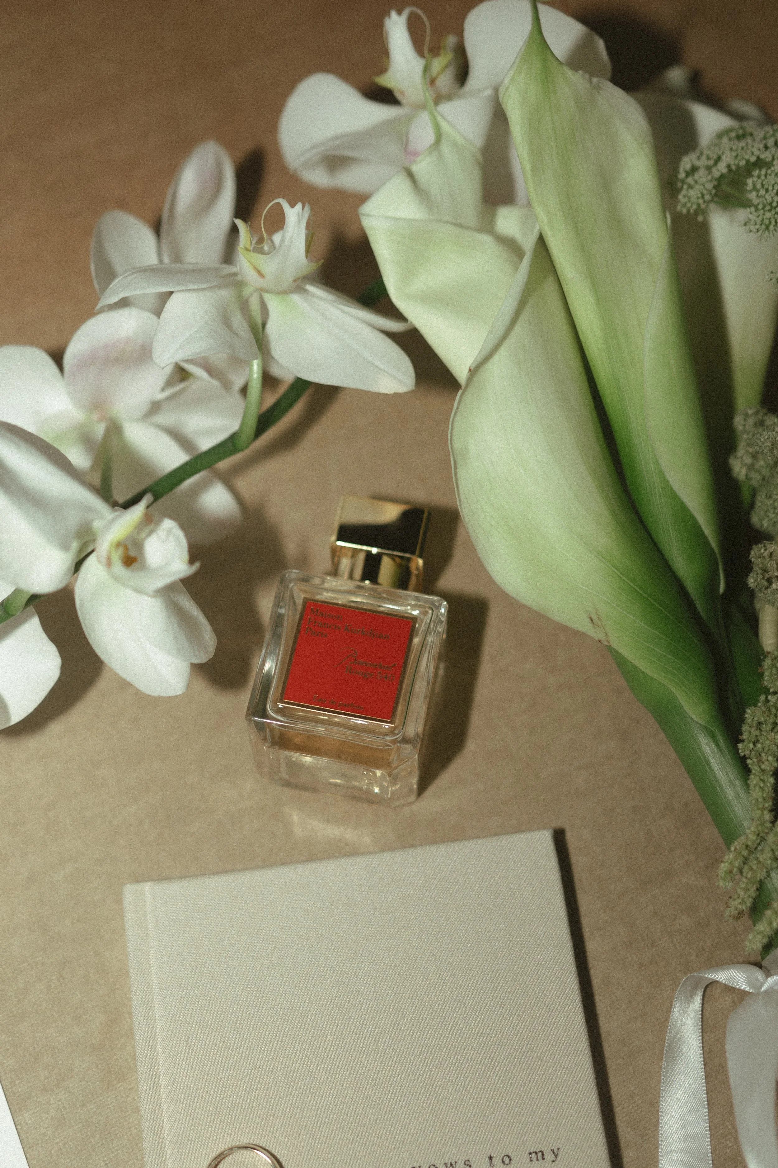 A bottle of perfume placed on a beige surface surrounded by white orchids, large green leaves, and a beige book or notebook.