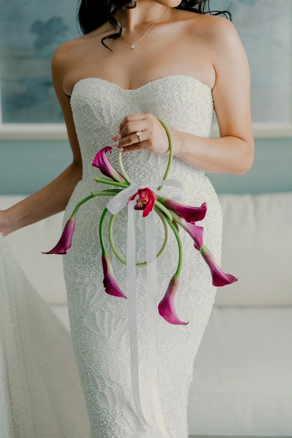A woman in a strapless white lace wedding dress holding a bouquet of purple calla lilies with a white ribbon, standing indoors.