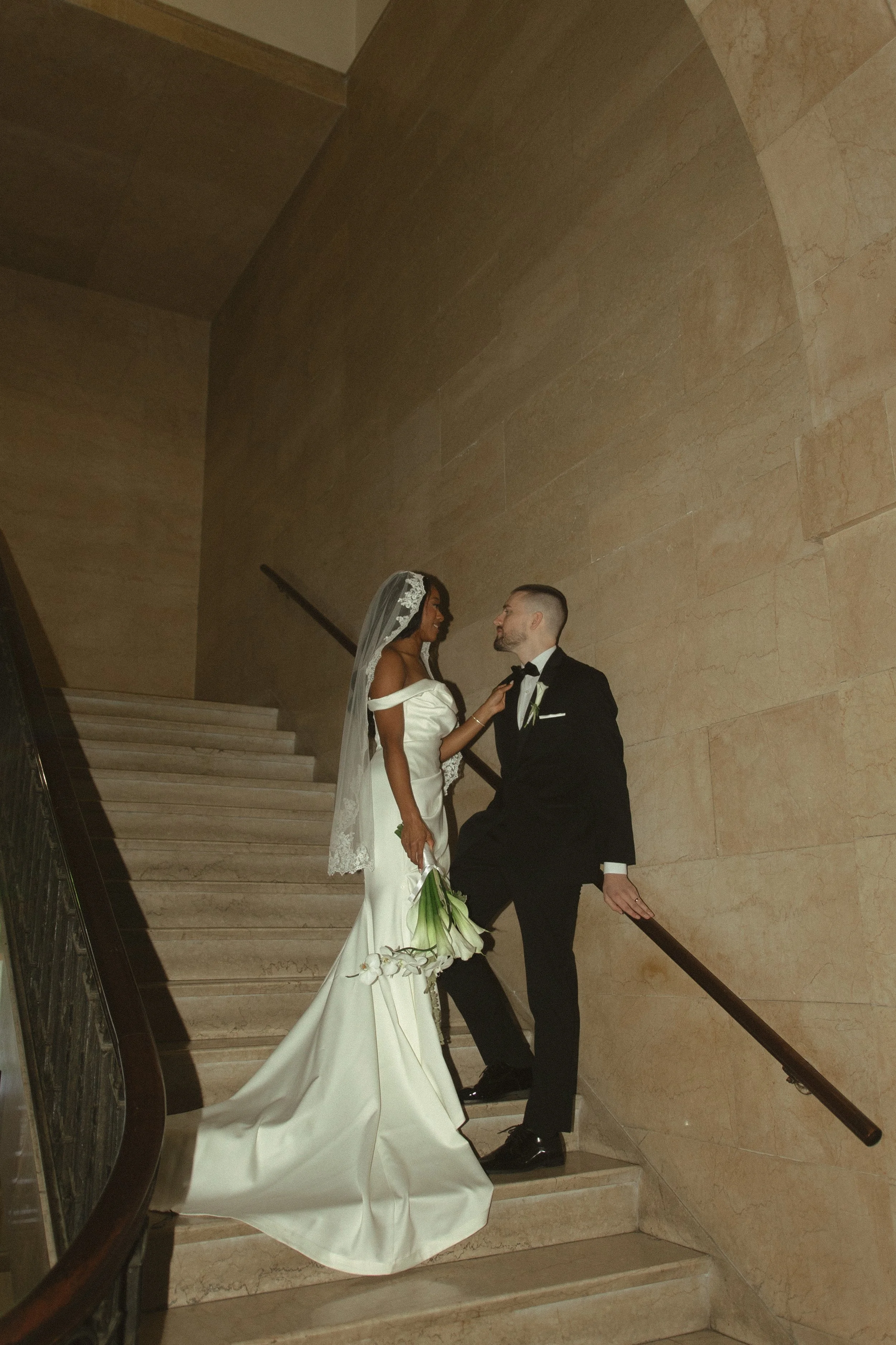 A bride and groom on a staircase, gazing at each other, with the bride holding a bouquet and wearing a wedding dress and veil, and the groom in a tuxedo.