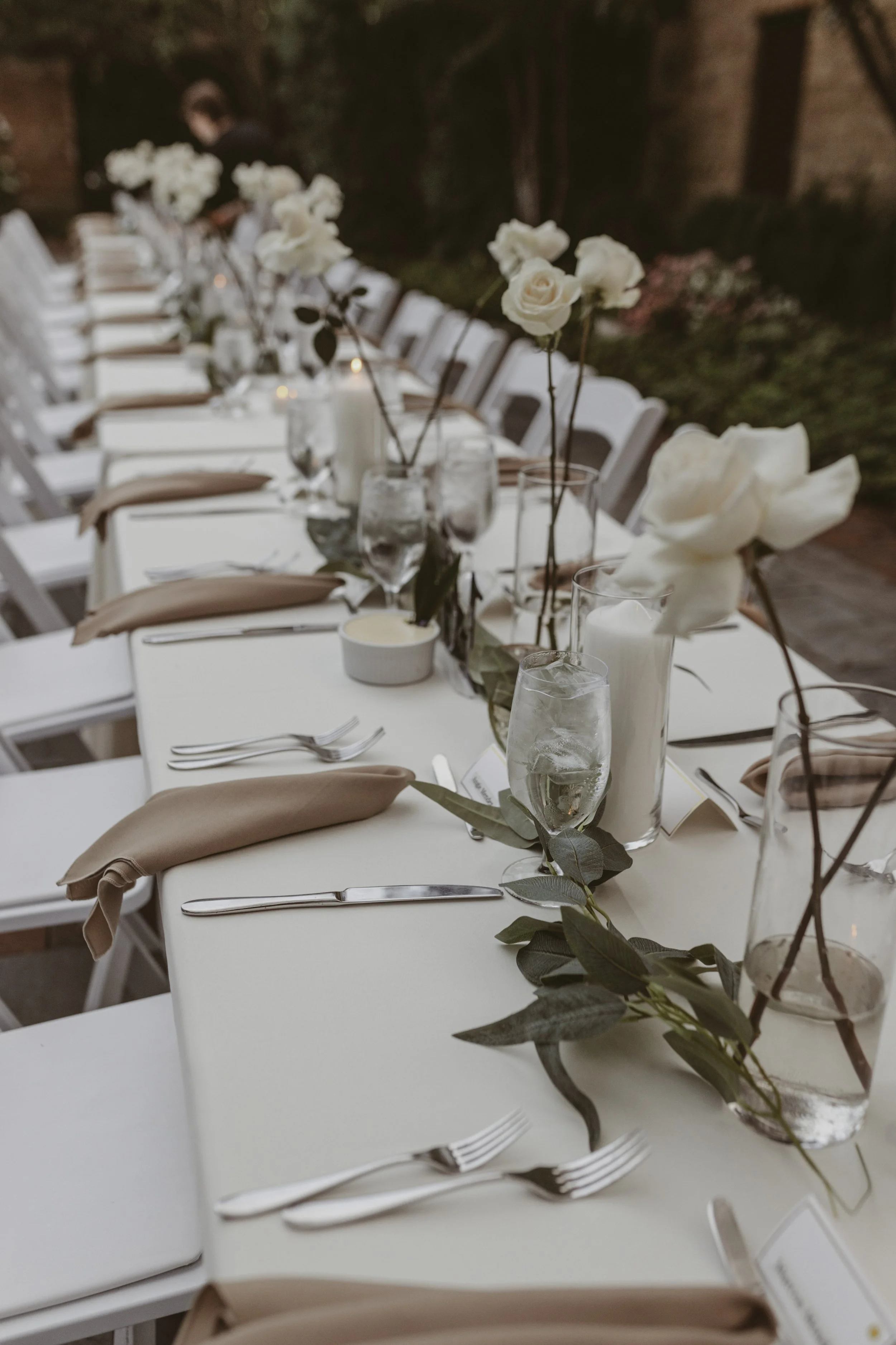 Elegant outdoor dining table set with white tablecloth, beige napkins, silverware, glassware, white roses, candles, and greenery, arranged for a formal event.