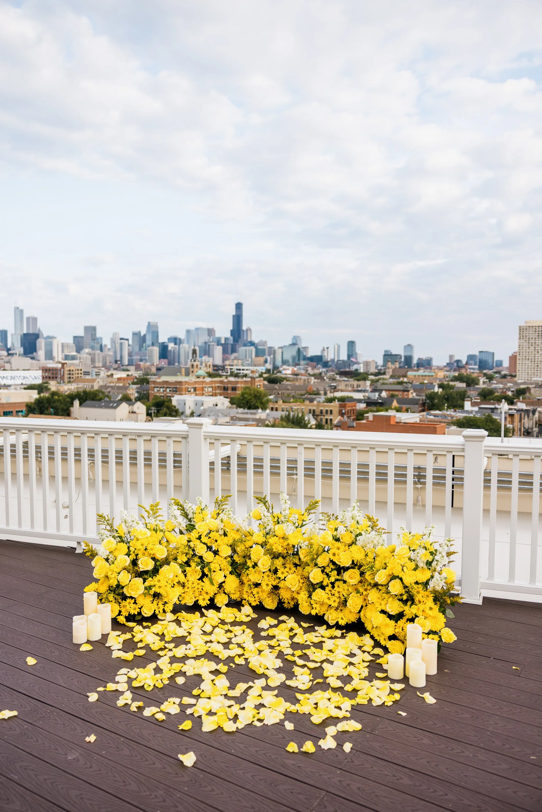 A rooftop with yellow flower arrangements, white candles, and rose petals against a city skyline background on a partly cloudy day.