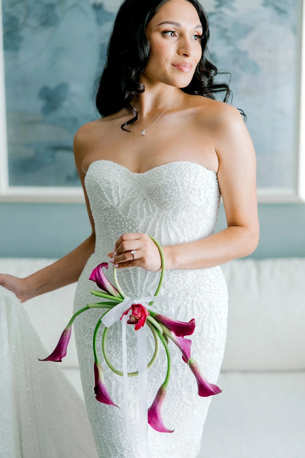 A woman in a strapless white wedding dress holding a bouquet of pink calla lilies, with dark wavy hair, wearing a necklace, and looking to the side near a gray wall with a framed artwork.
