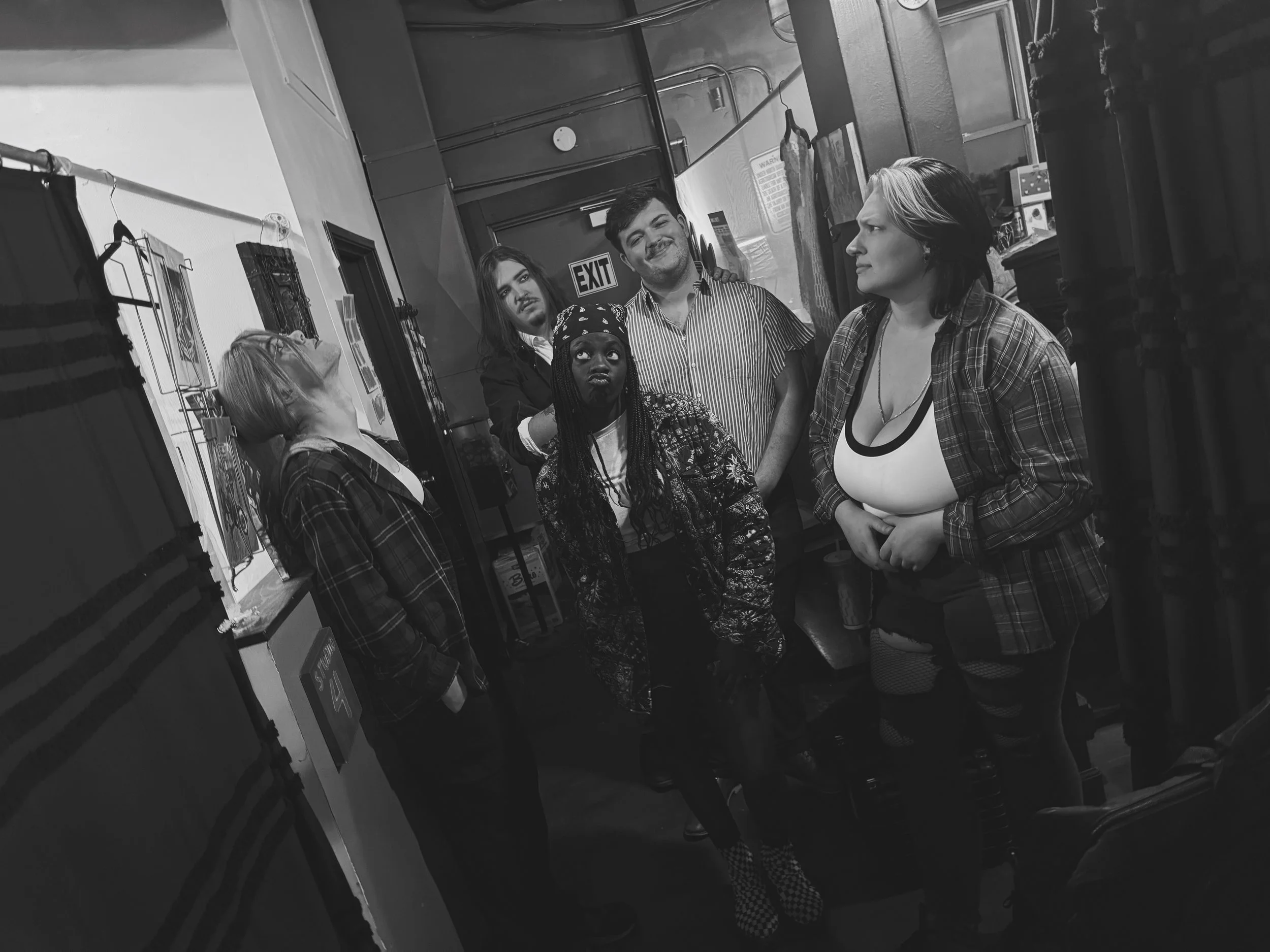 A group of six diverse young people standing together in what appears to be an indoor setting, possibly a gallery or studio, with artwork and display racks around them. They are engaged in conversation, with some looking at each other and one looking upward.