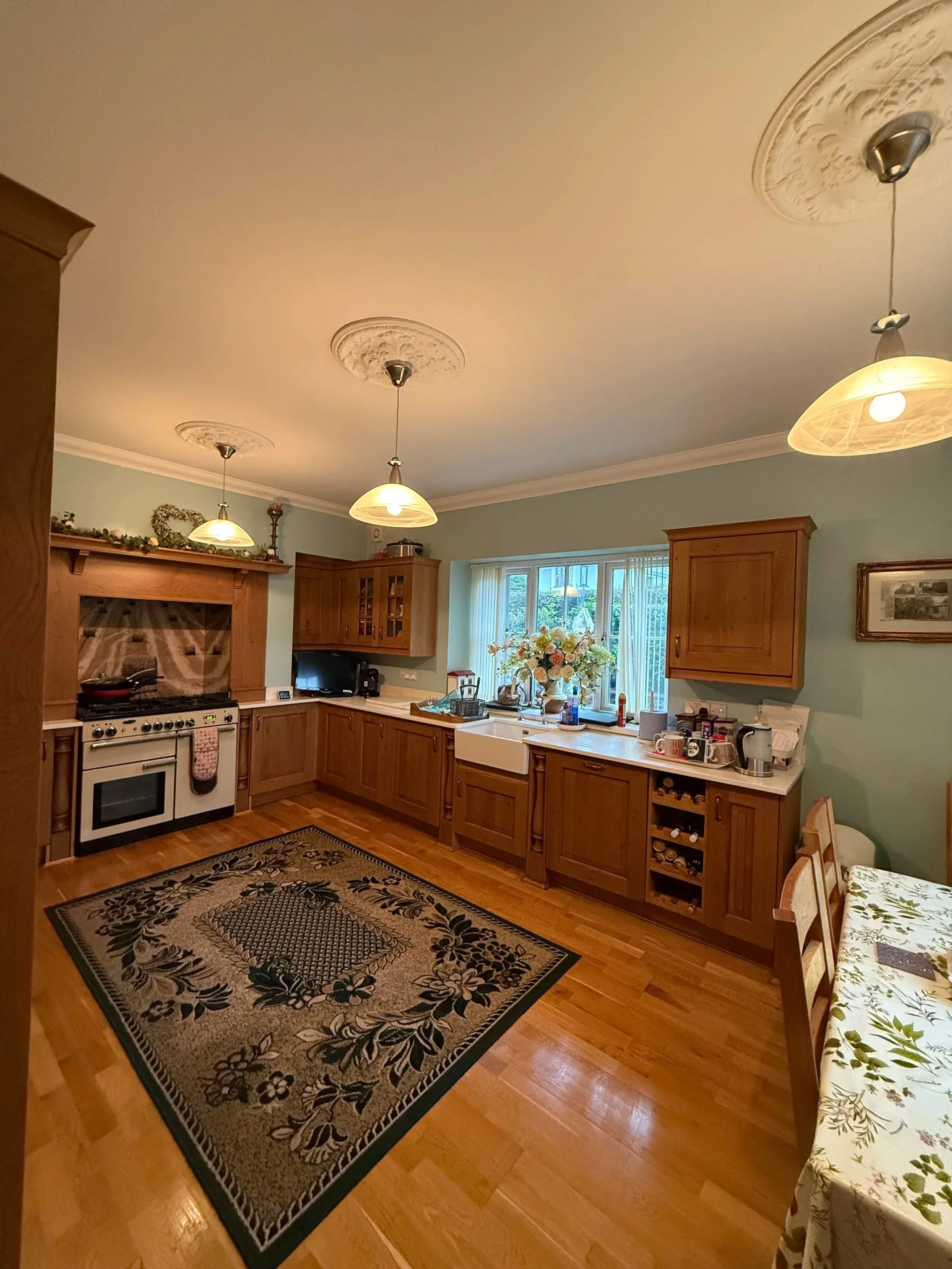 A cozy kitchen with wooden cabinets, a central window with a flower vase, a stove, and a dining table with a floral tablecloth.