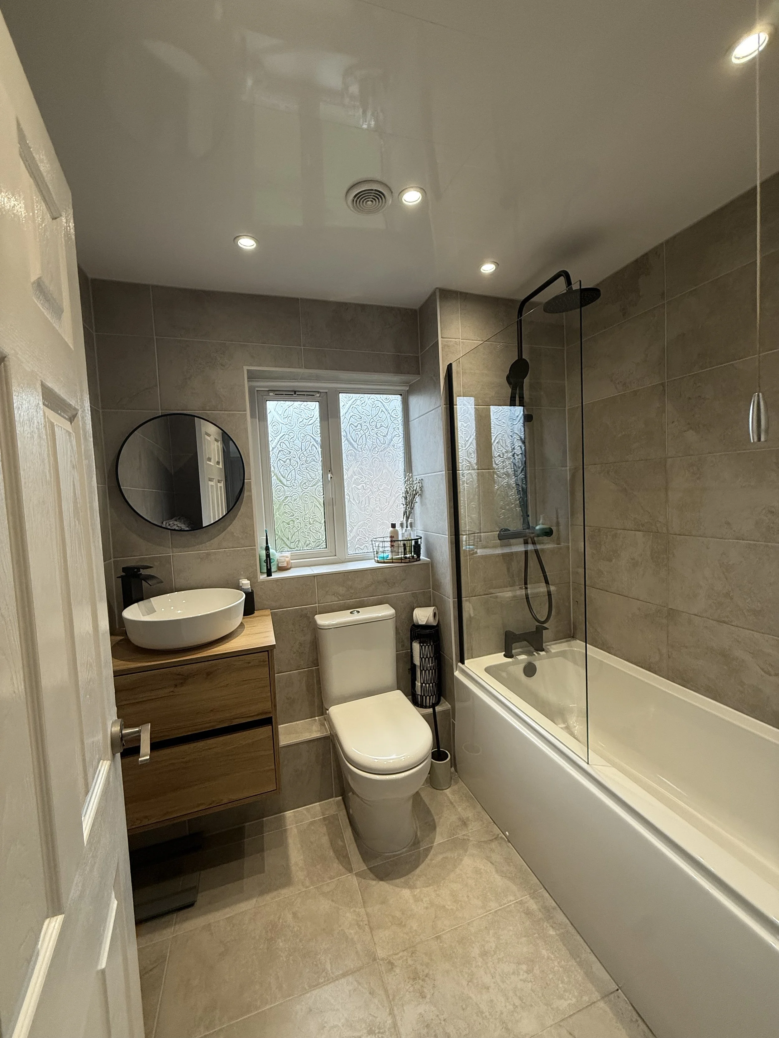 Modern bathroom with beige tiled walls and floor, a bathtub with a glass shower screen, a black overhead shower, a white toilet, a wooden vanity with a vessel sink, a round mirror, a window, and ceiling lights.