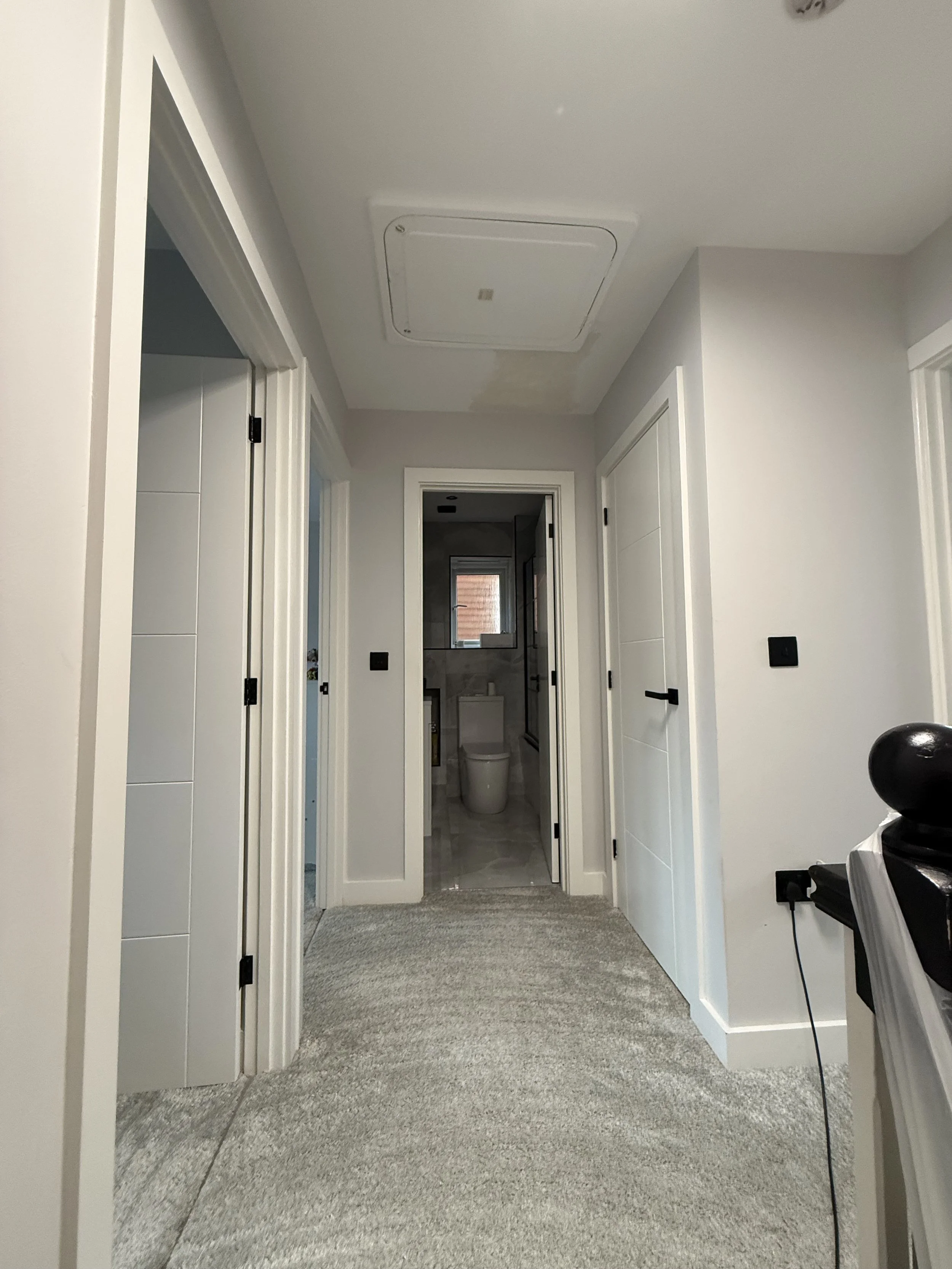 A hallway in a house with three doors, leading to a bathroom at the end. The floor is carpeted with a light gray rug, and the walls are painted white. There is a ceiling access panel and a vent on the ceiling.